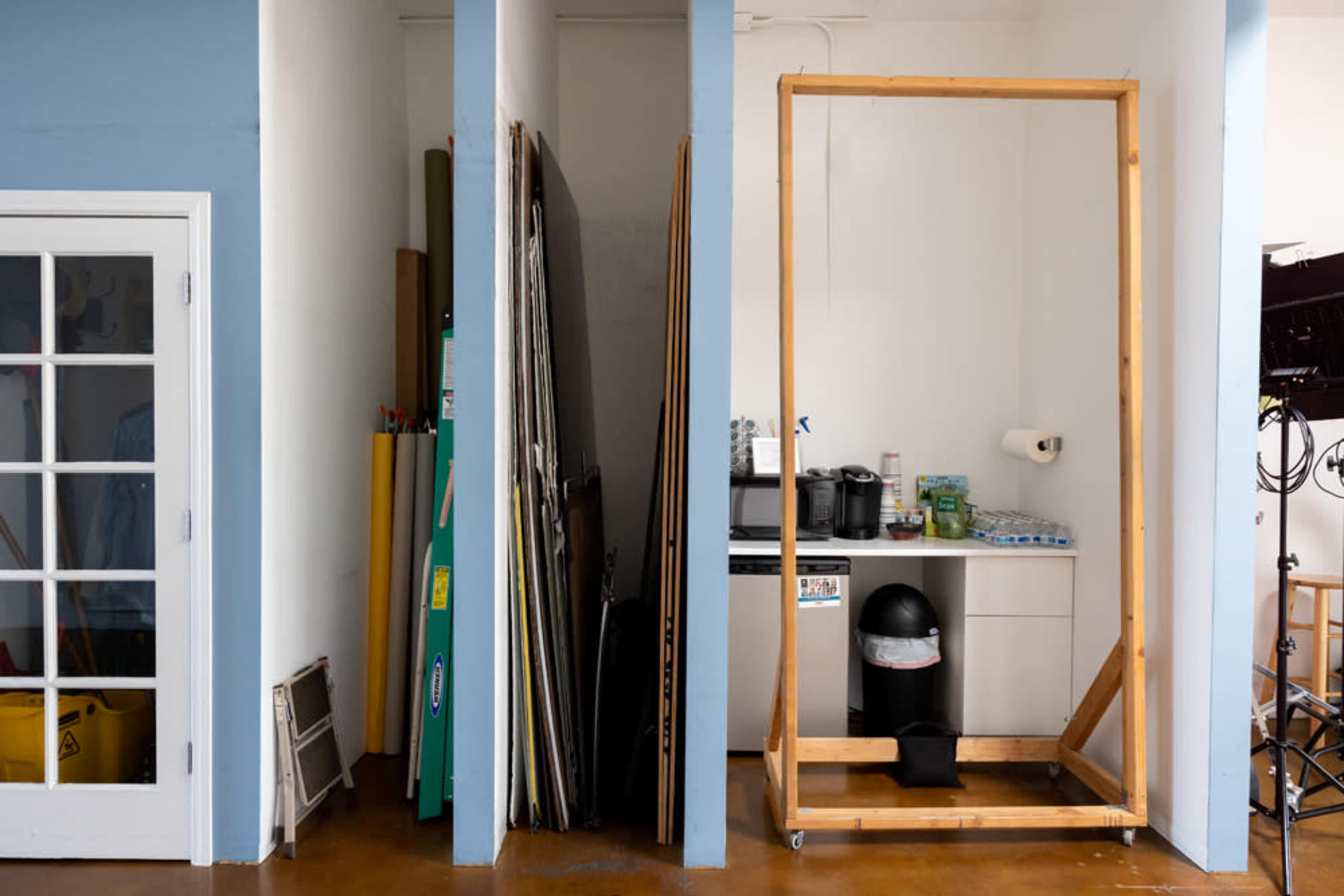 A minimalist studio space features stacked boards and a large wooden frame beside a kitchenette area with a trash bin and supplies.