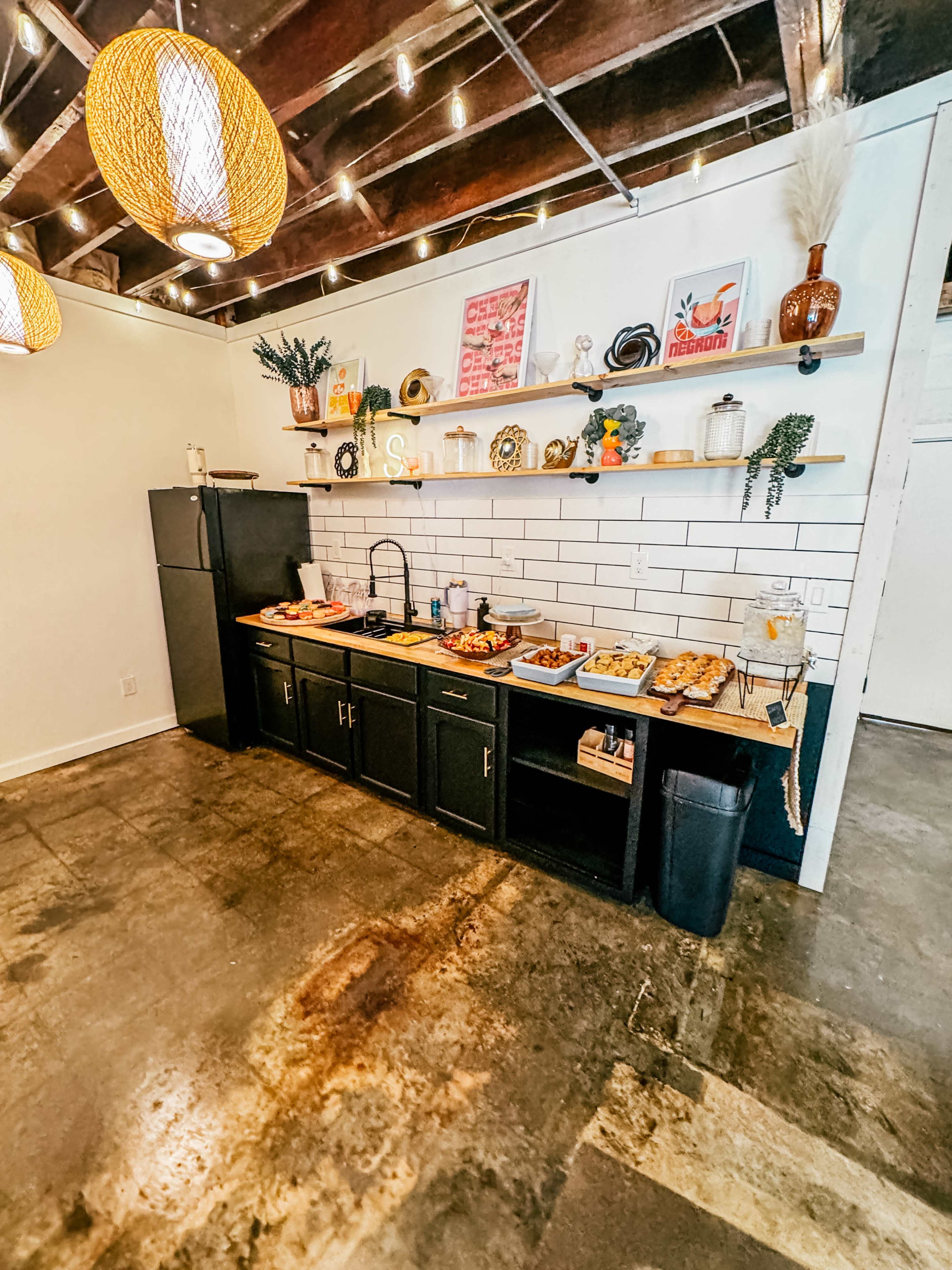 A kitchen area with a black countertop and various food items neatly arranged on it, against a backdrop of white tiles and decorative shelves.