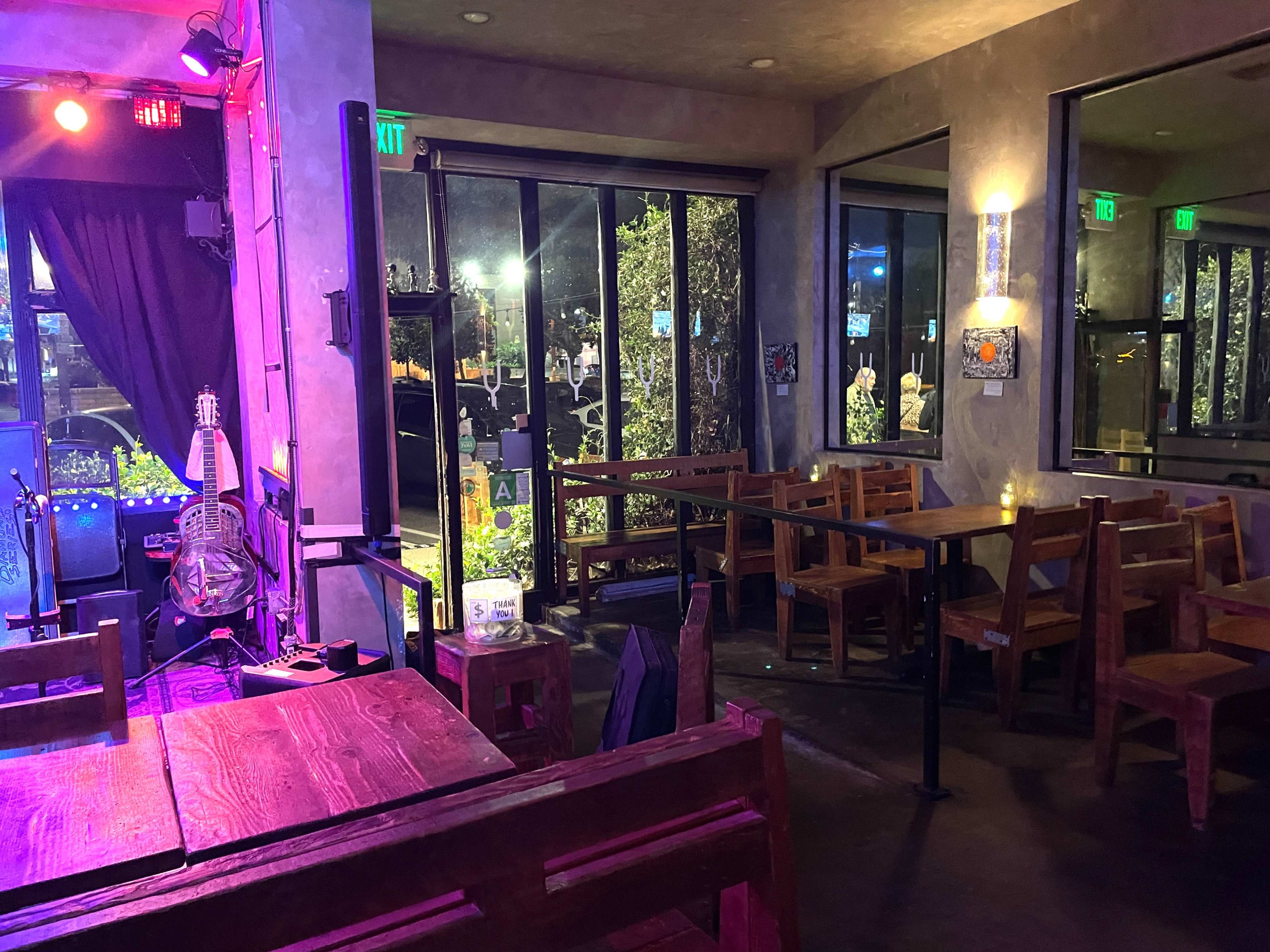 The image shows a dimly lit restaurant interior with wooden tables and chairs, large windows revealing an outdoor scene, and stage lighting creating an atmospheric effect.