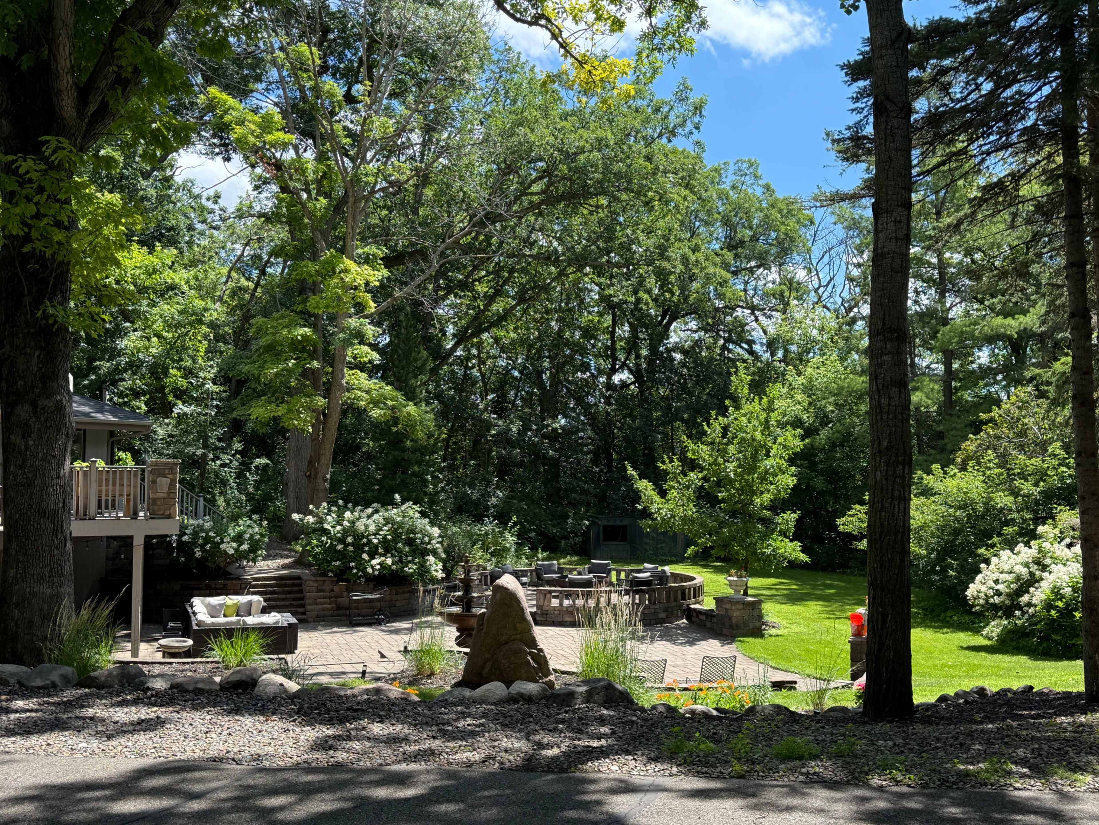 The image shows a landscaped outdoor area featuring a circular stone patio surrounded by greenery, trees, and a clear blue sky.