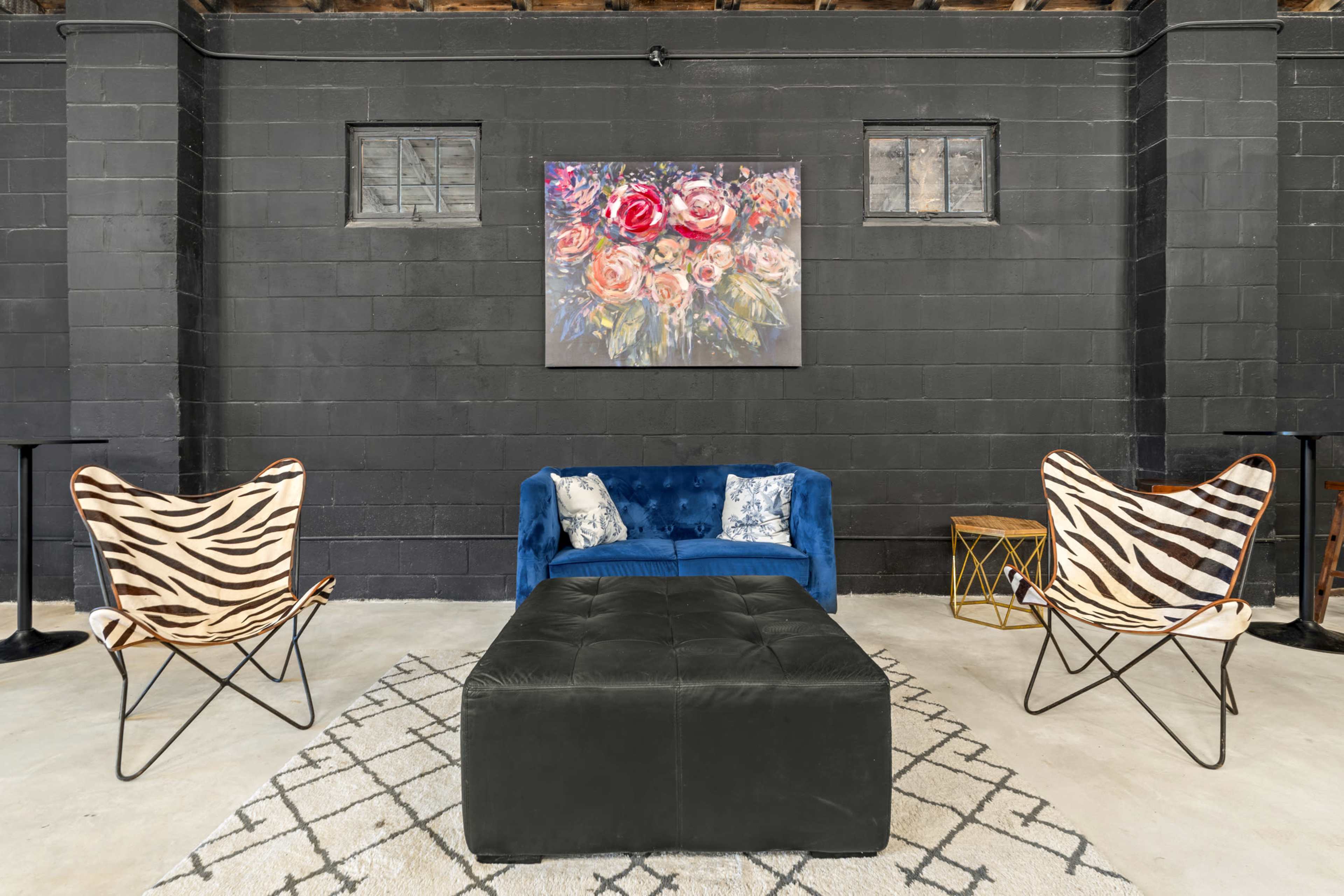 A blue couch, two patterned chairs, and an ottoman are arranged on a rug in a room with black walls and a floral painting above.