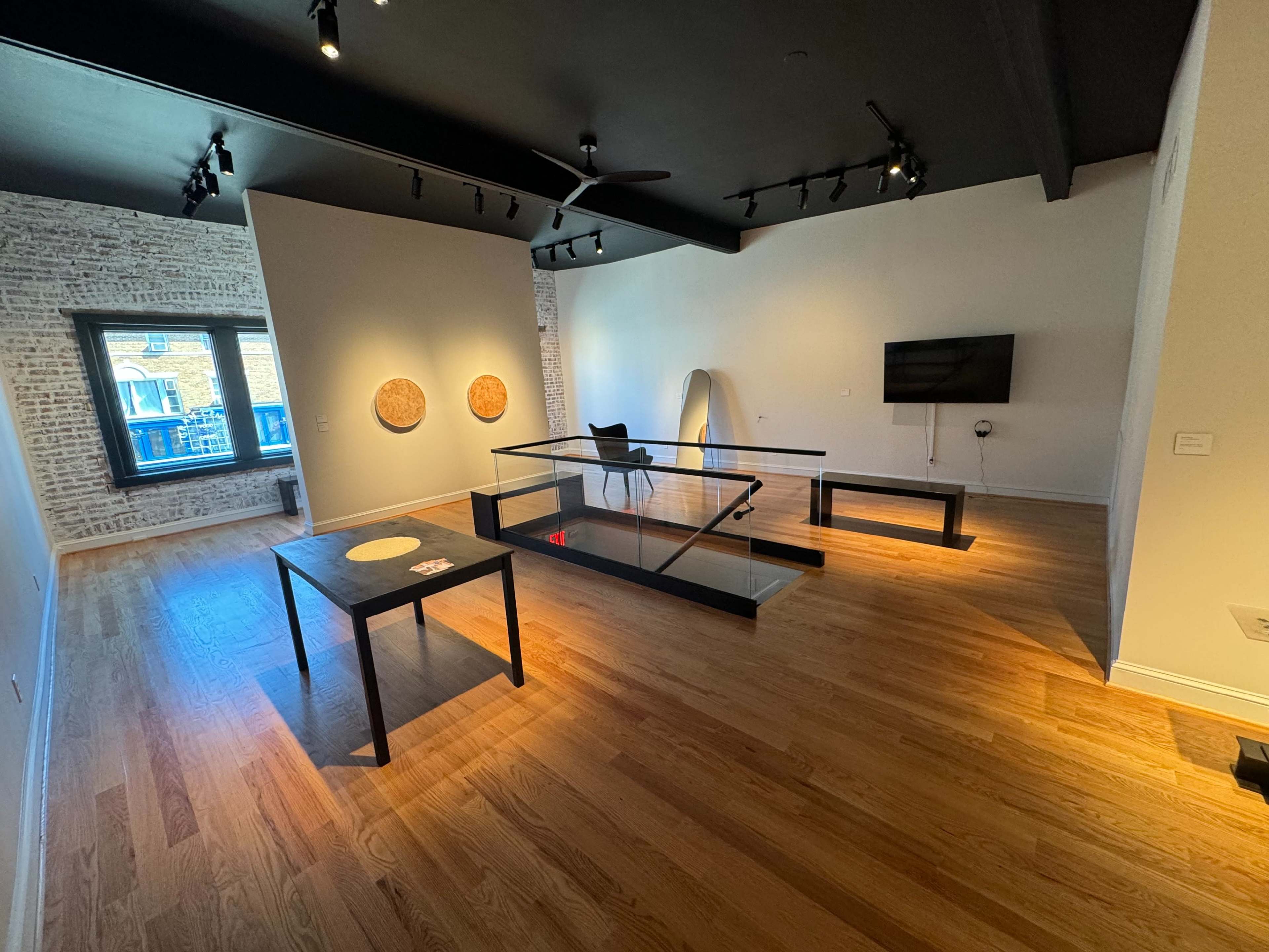 A spacious, modern room with wooden flooring, featuring a large glass table, a chair, and two circular artworks on the walls.