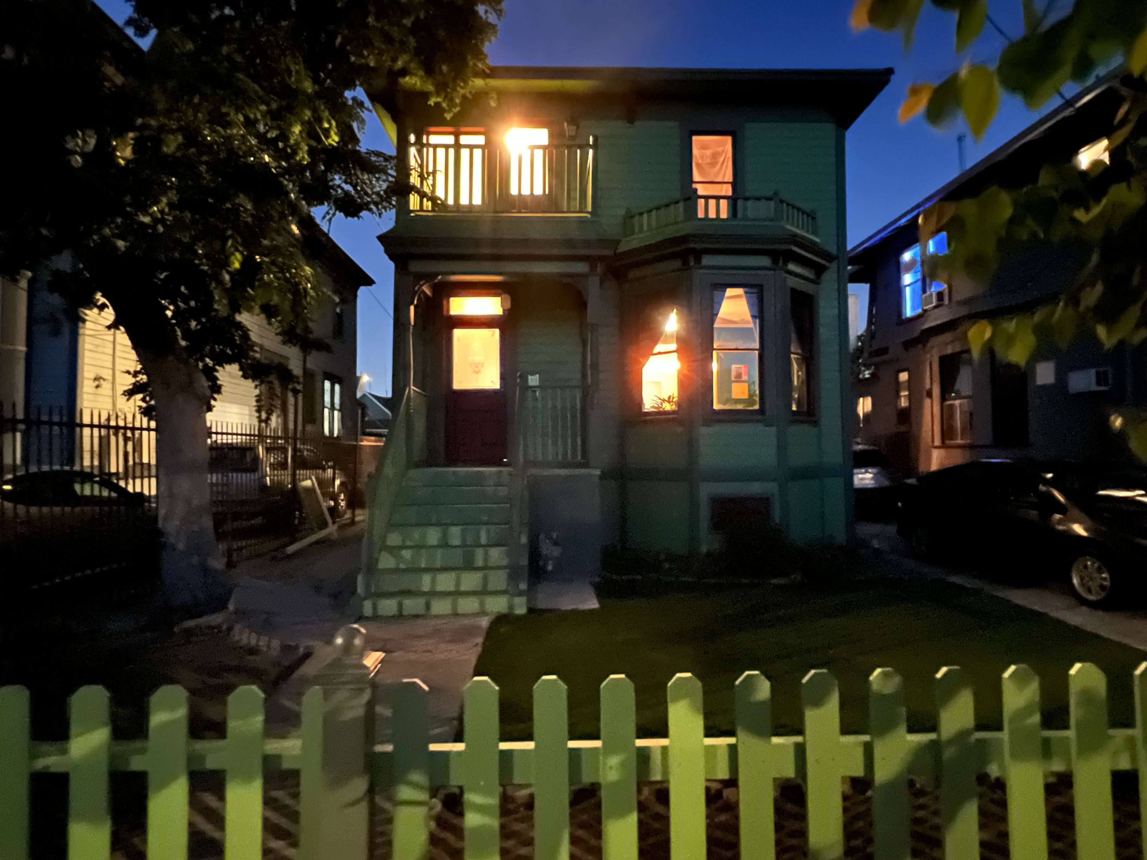A two-story green house with a staircase and a porch is illuminated by light from the windows at dusk, surrounded by a white picket fence.