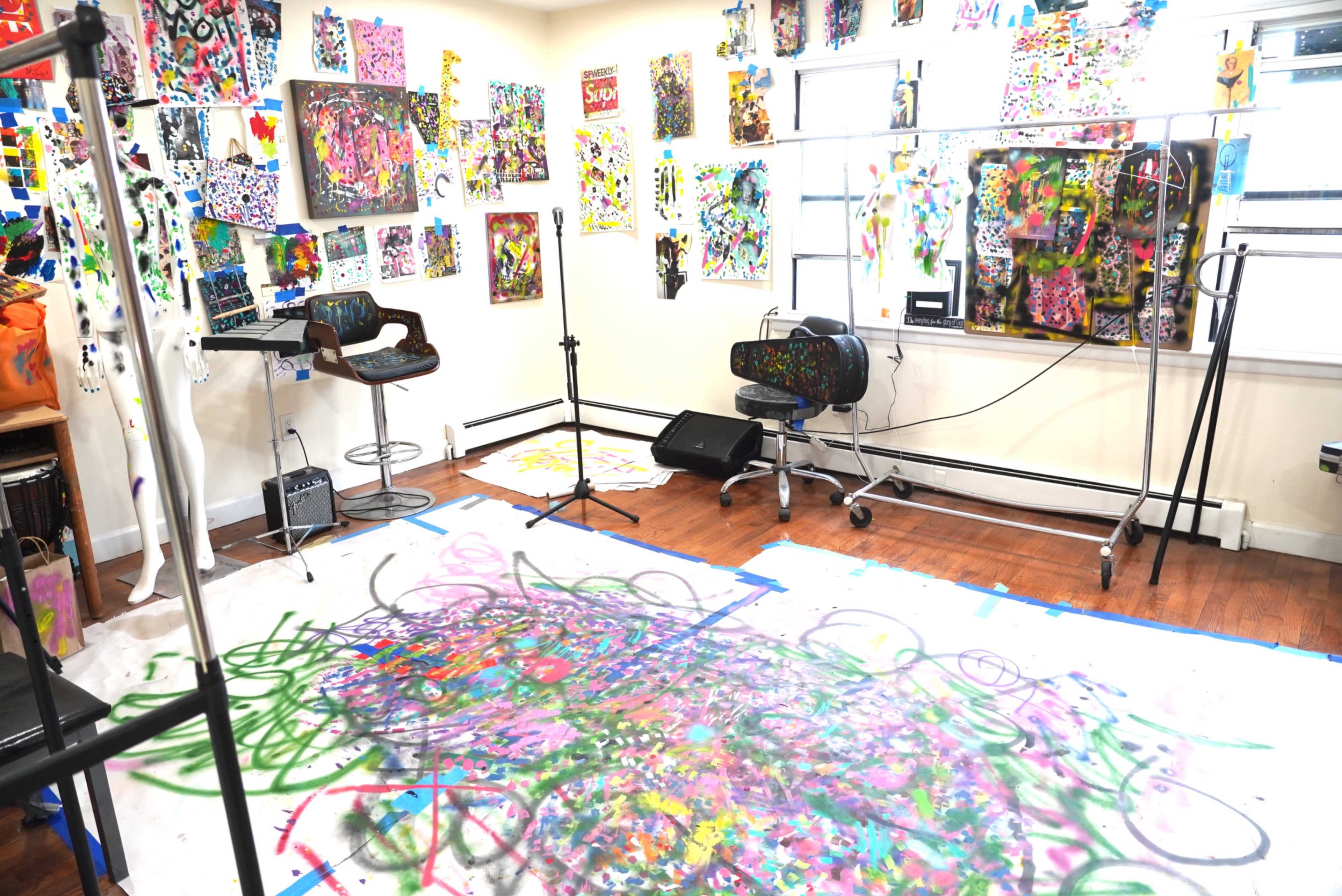 The image shows a vibrant art studio filled with colorful paintings on the walls and a paint-splattered floor.