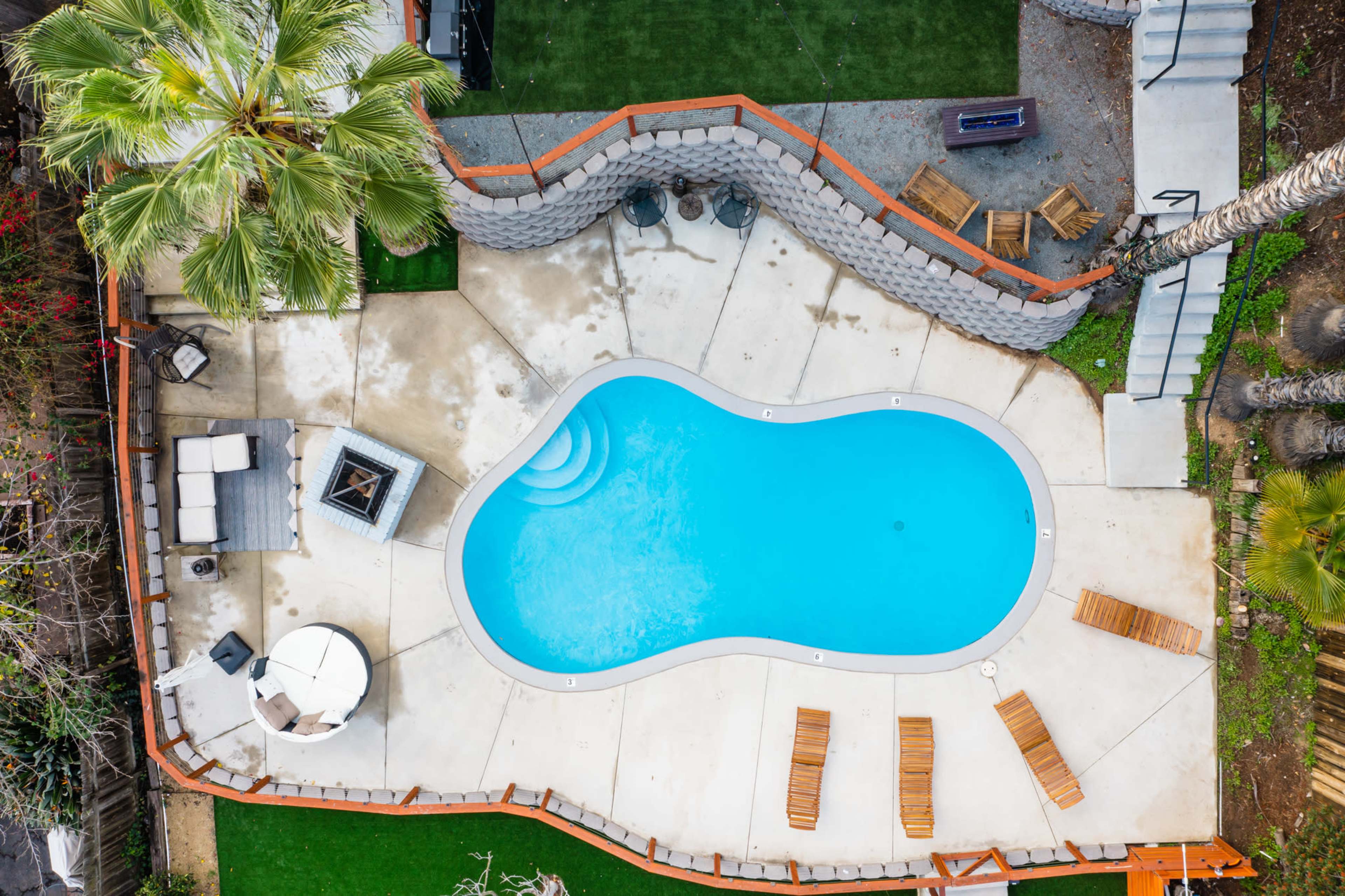 An aerial view shows an irregularly shaped swimming pool surrounded by concrete, lounge chairs, a fire pit, and decorative landscaping.