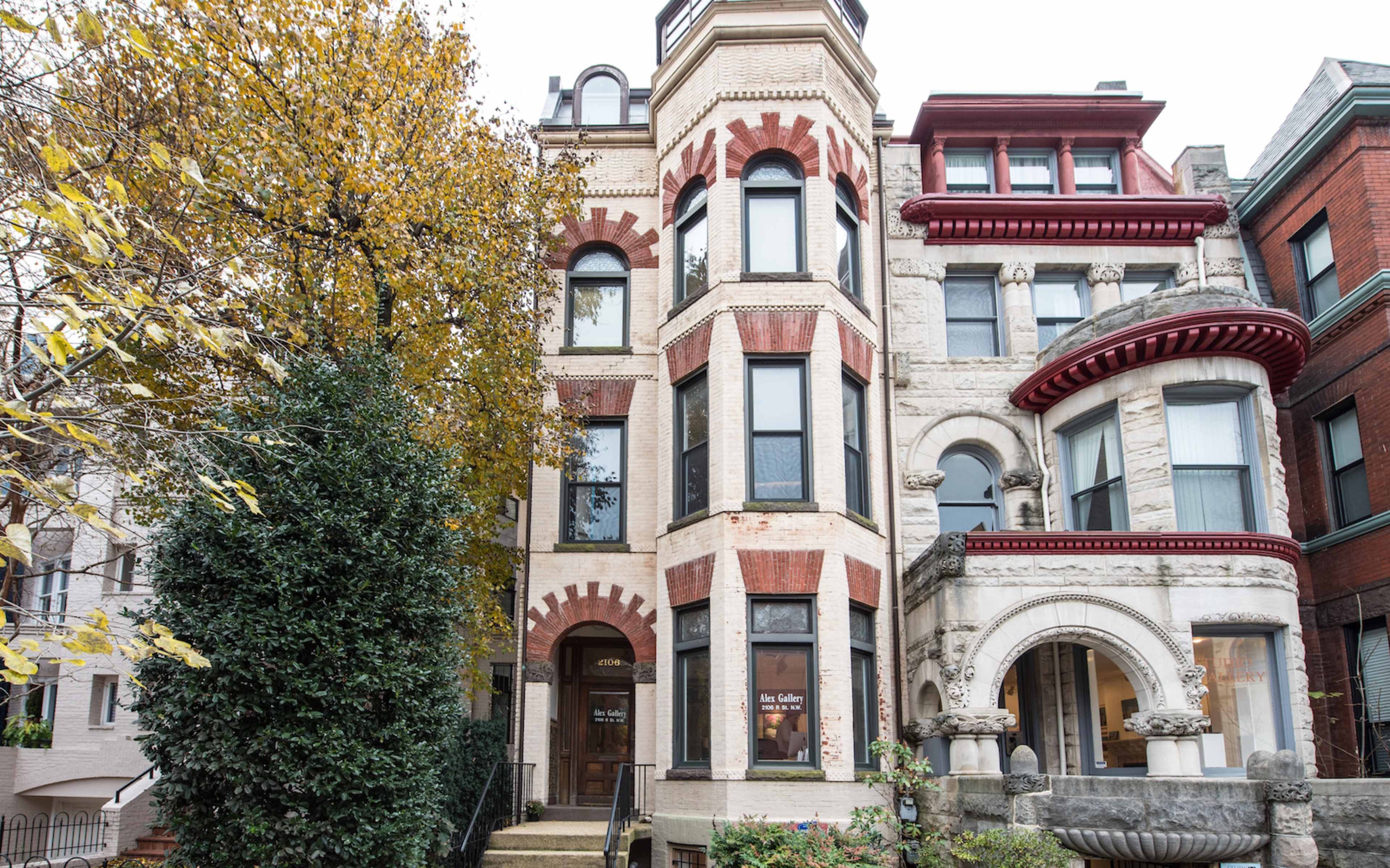 The image shows a historic multi-story building with intricate architectural details, featuring a mix of red brick and stone, flanked by trees and neighboring structures.