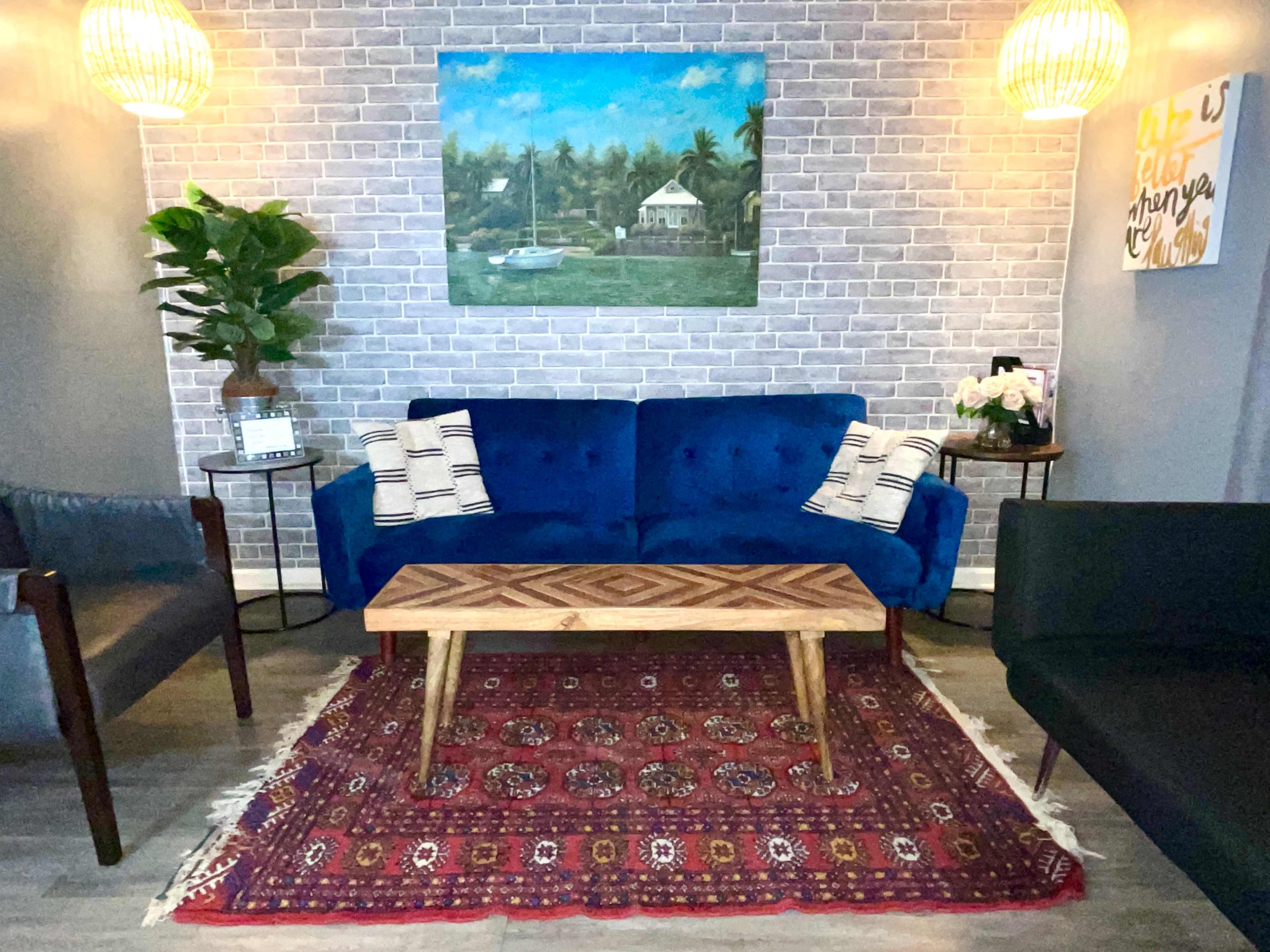The scene features a living area with a blue couch, two black chairs, a wooden coffee table on a patterned rug, and decor including a painting and potted plants against a brick wall.