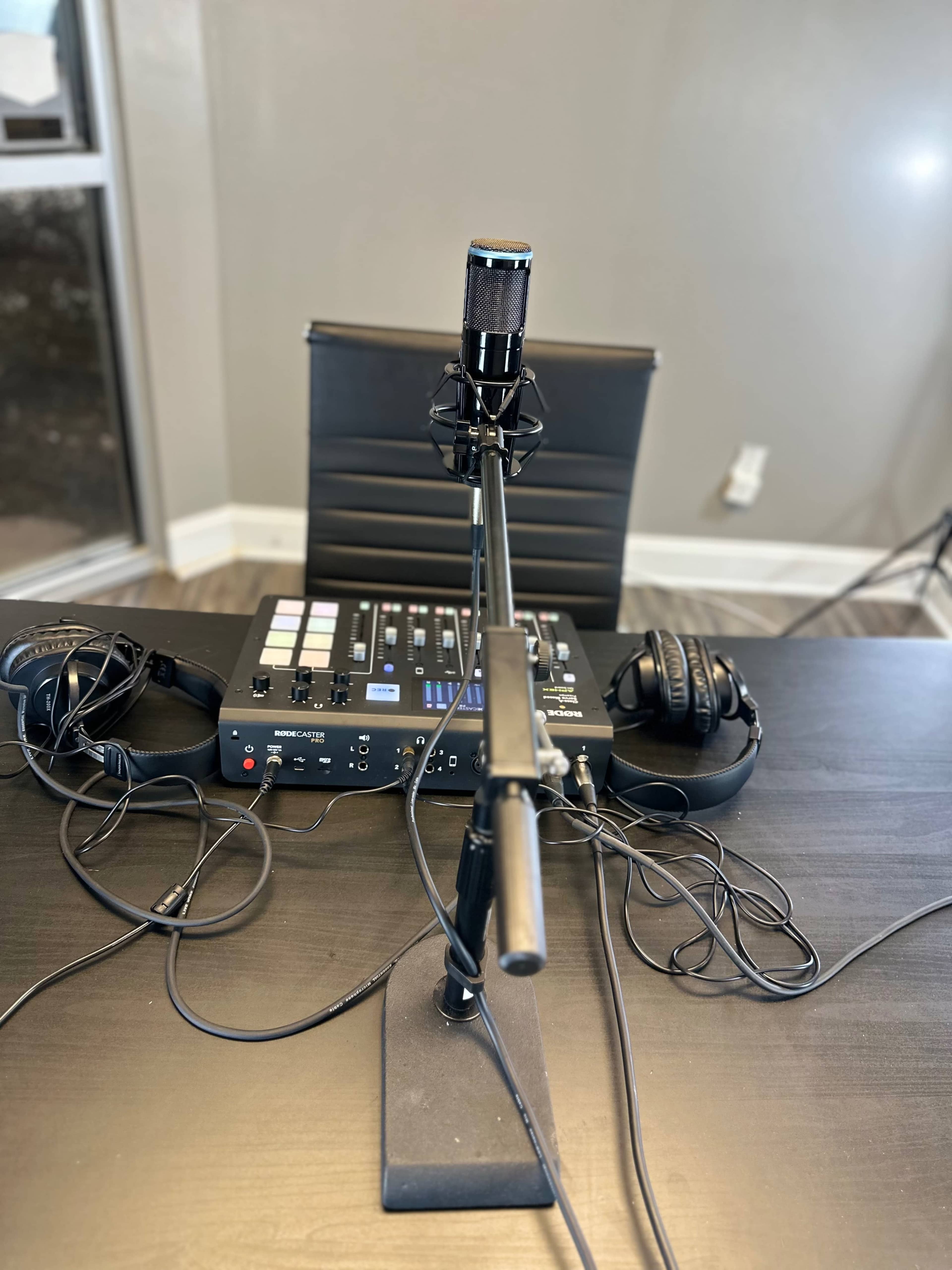 A microphone is mounted on a stand in front of a mixing console and headphones on a black table.