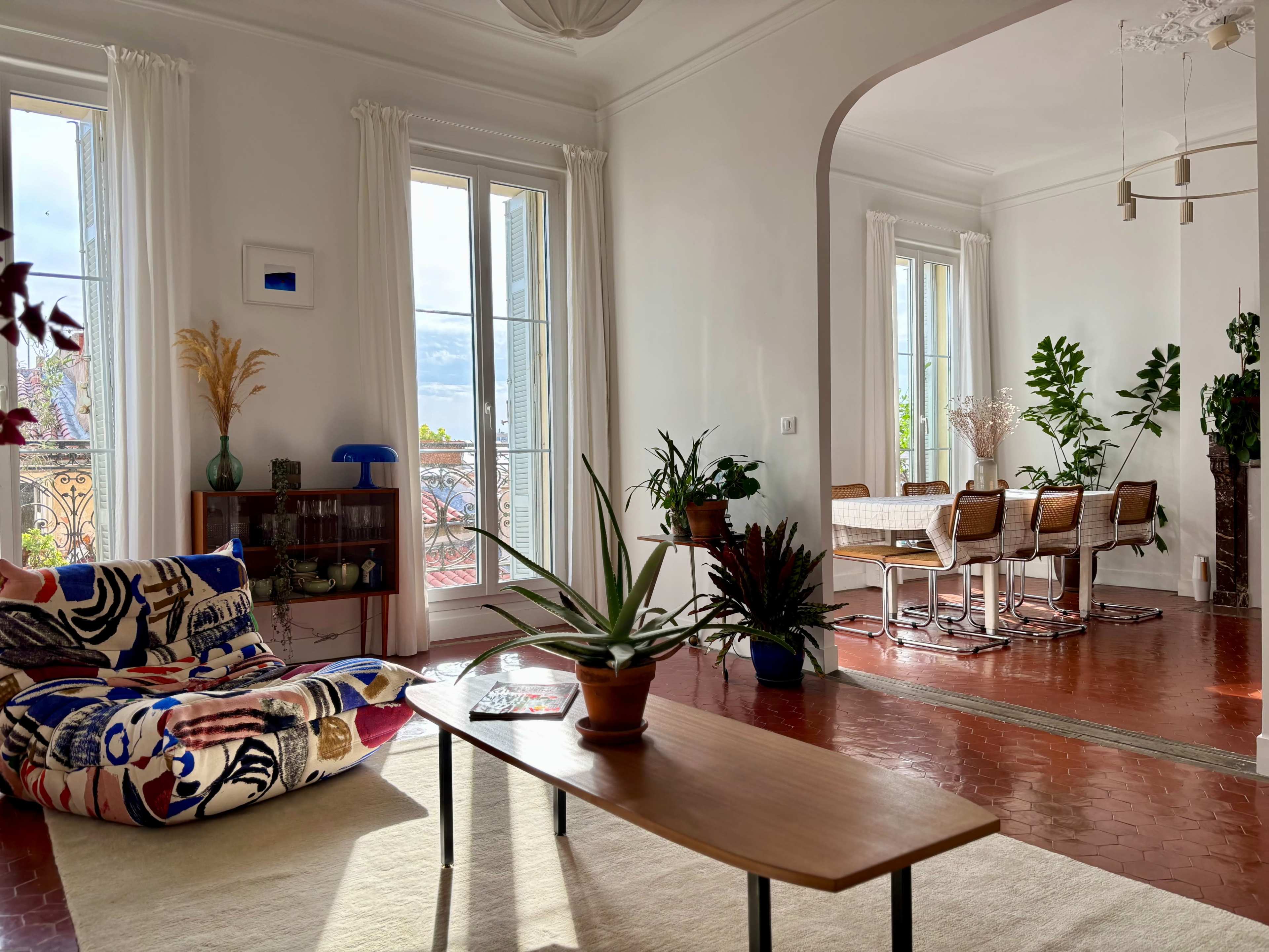 A spacious living room features a colorful patterned chair and a wooden coffee table with plants, leading to a dining area with a table and chairs near large windows.