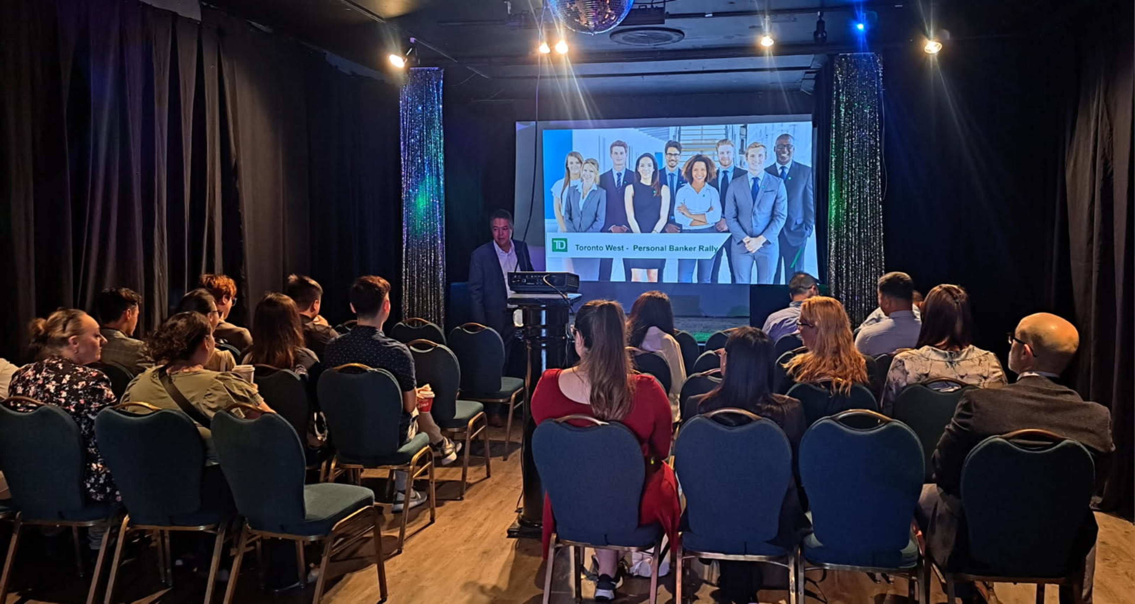 A speaker presents to an audience in a dimly lit venue featuring a large screen displaying a group photo of professionals.