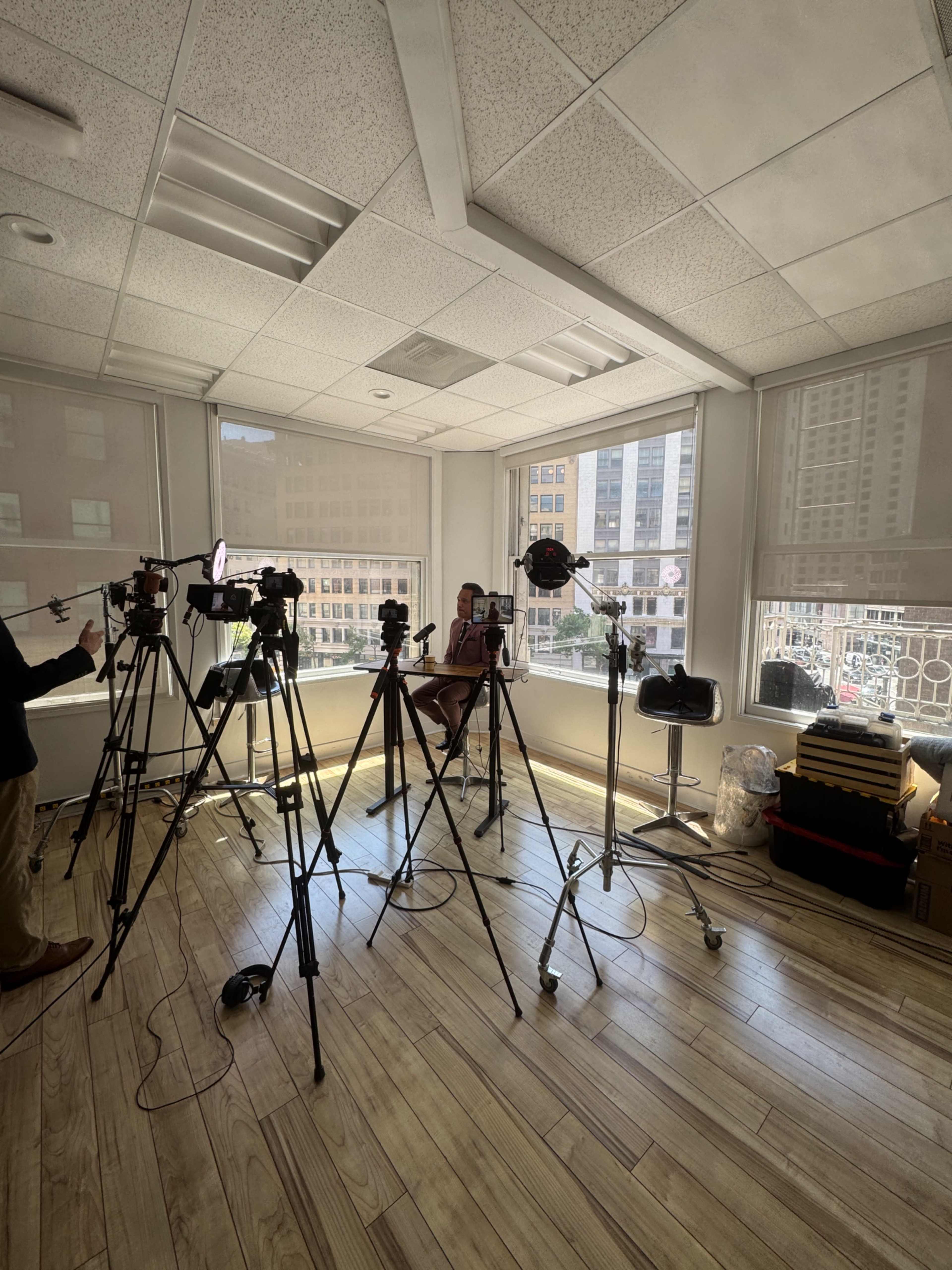 A well-lit room with large windows contains multiple camera stands and lighting equipment arranged around a seated subject.