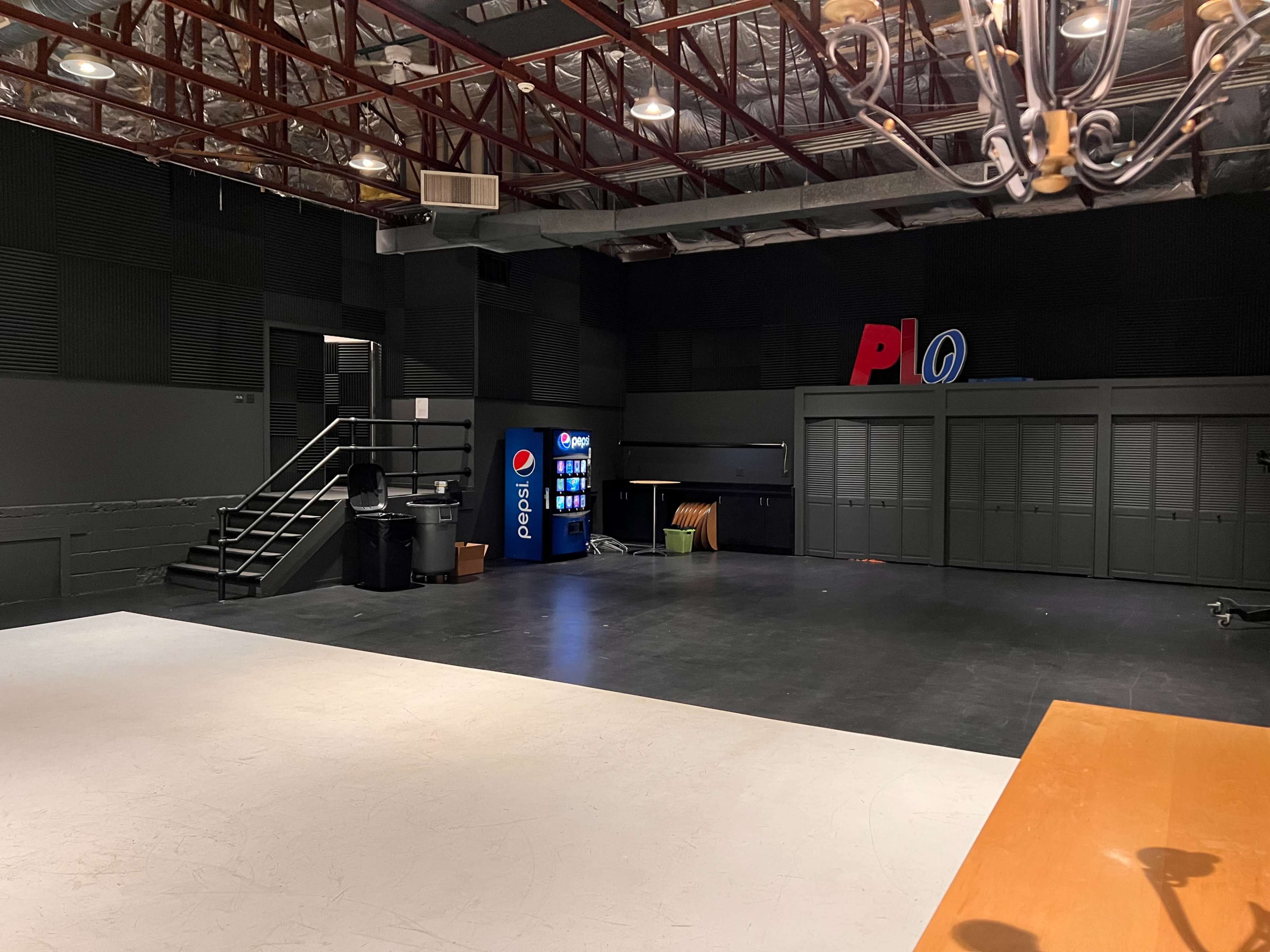 A spacious, dimly lit room with a Pepsi vending machine, a staircase, and a set of closed cabinets along one wall.
