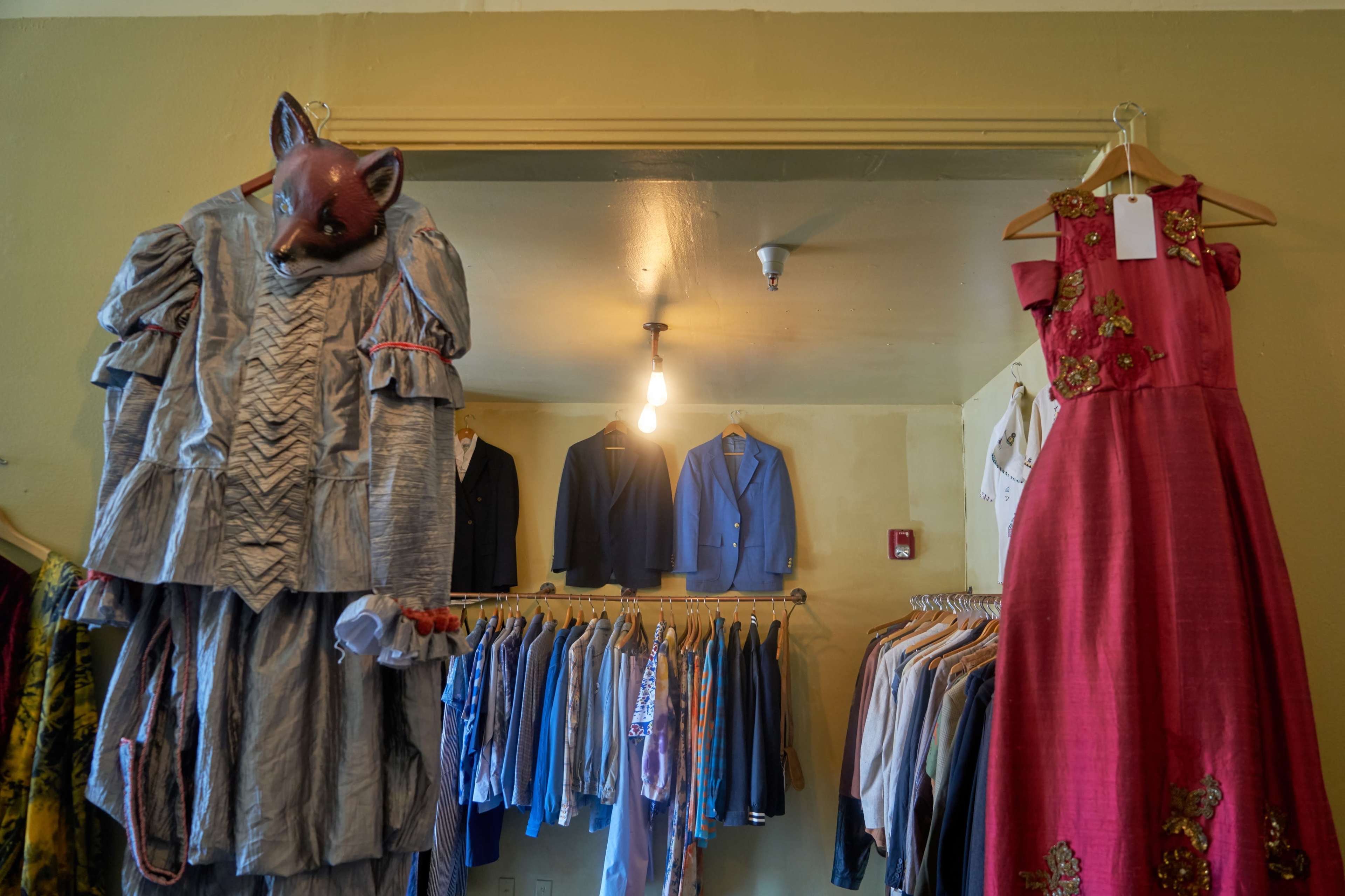 A vintage clothing store displays a variety of garments, including a costume with a fox mask and formal suits, arranged on hangers.
