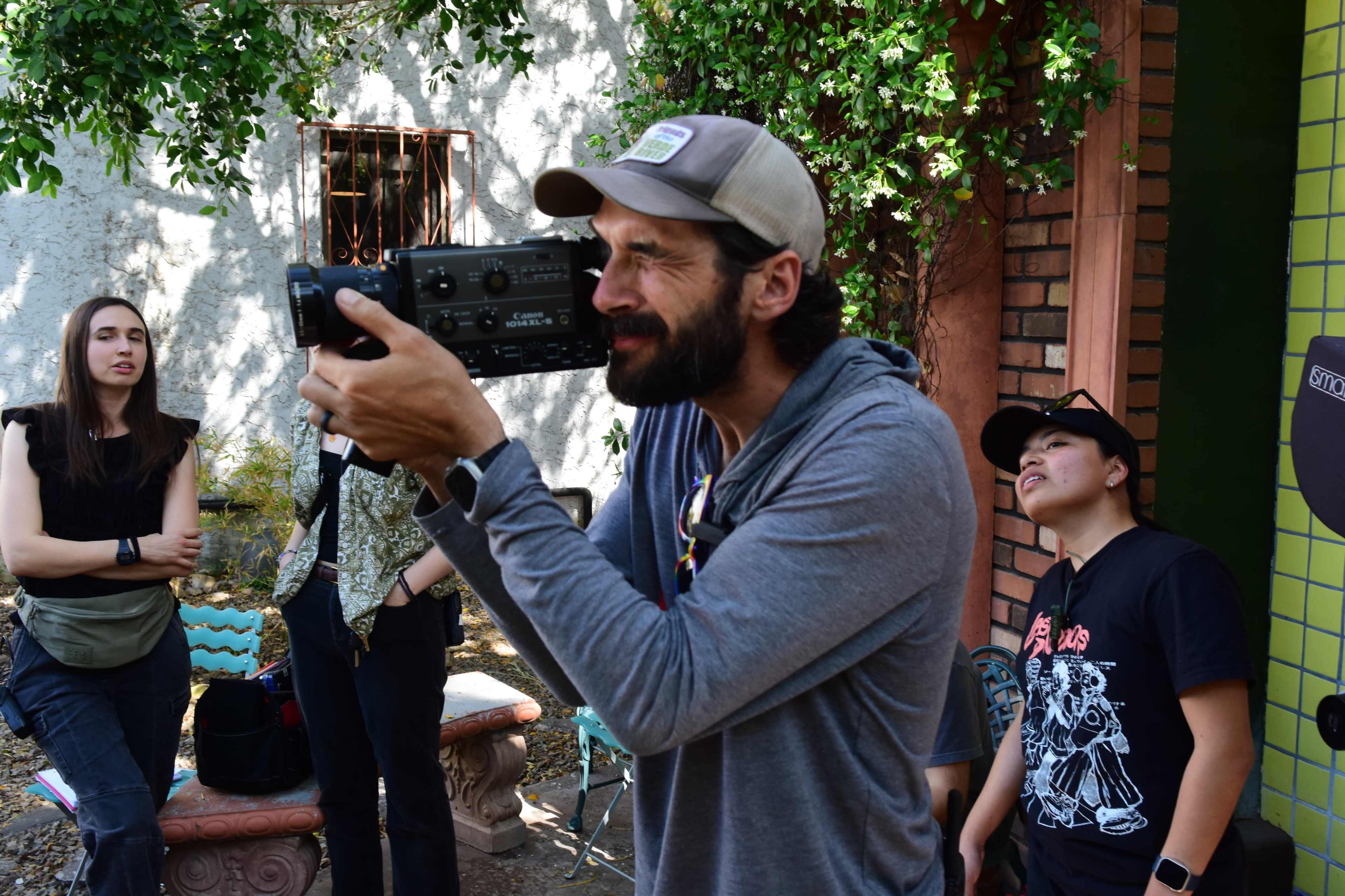A man holds a video camera while three individuals stand nearby in a garden setting.