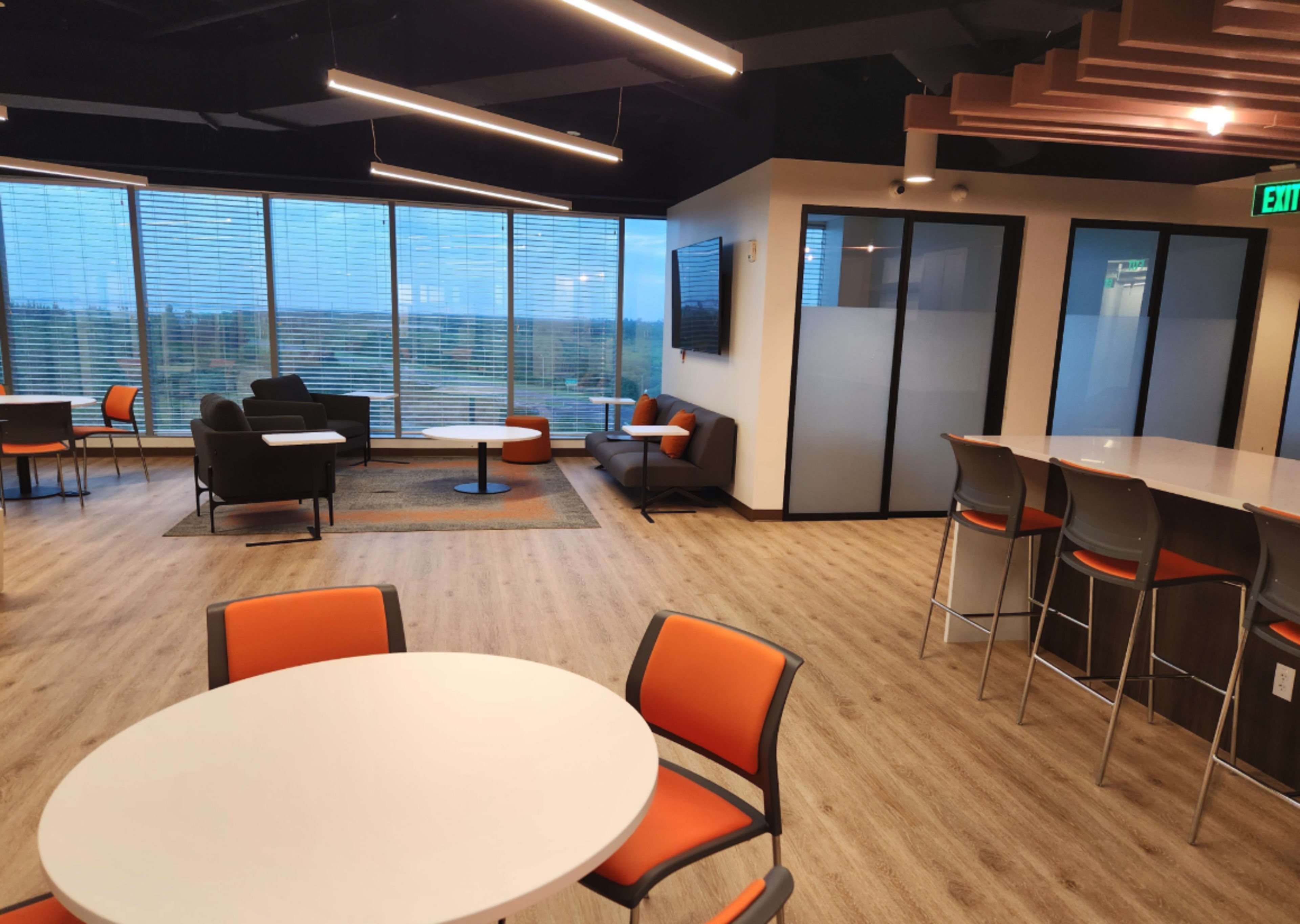 The image shows a modern office lounge area with tables, chairs, and a wall of windows displaying an outdoor view.