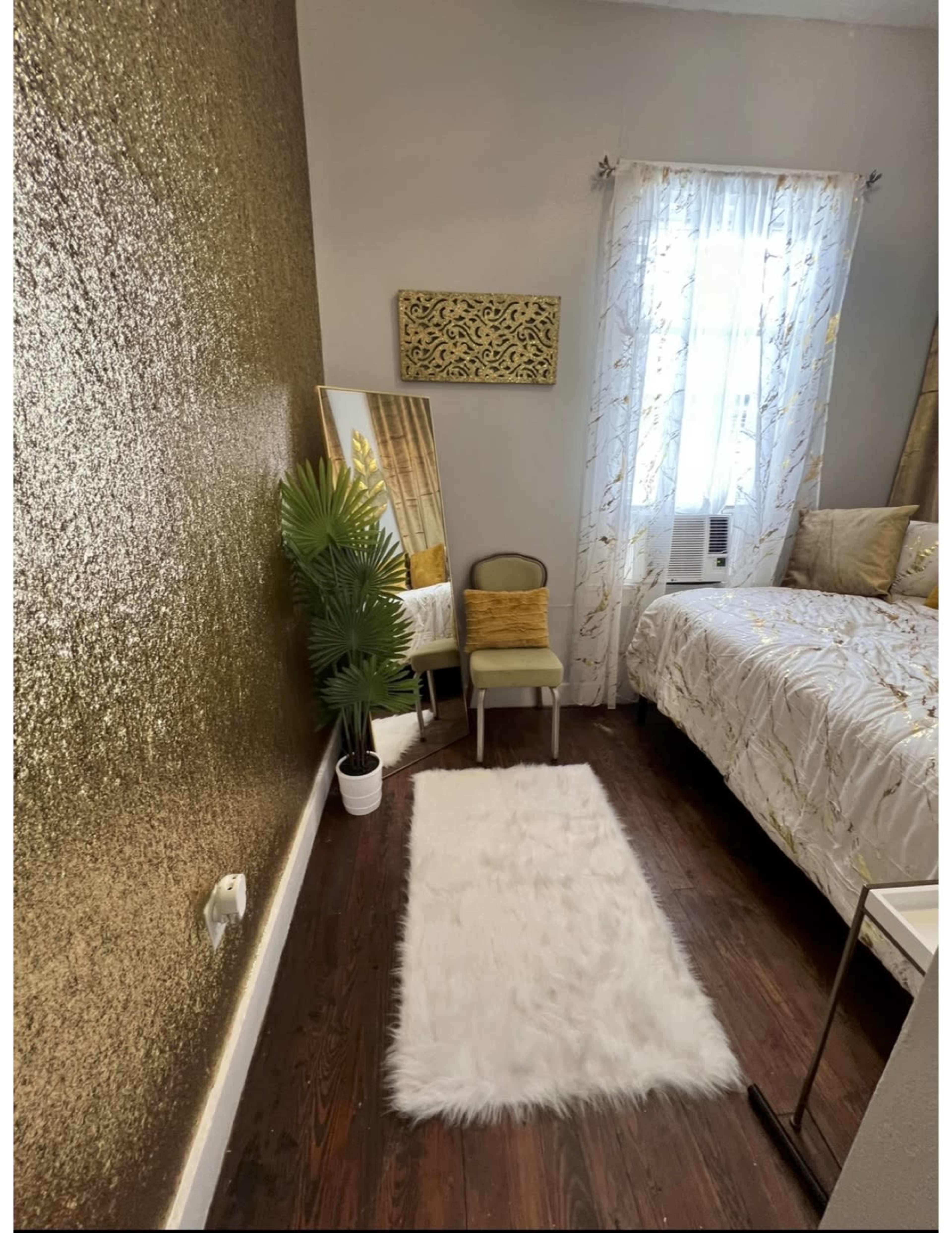A bedroom with a gold-accented wall, a white rug on the dark wooden floor, a small chair with a cushion, a mirrored reflection, and a window with sheer curtains.