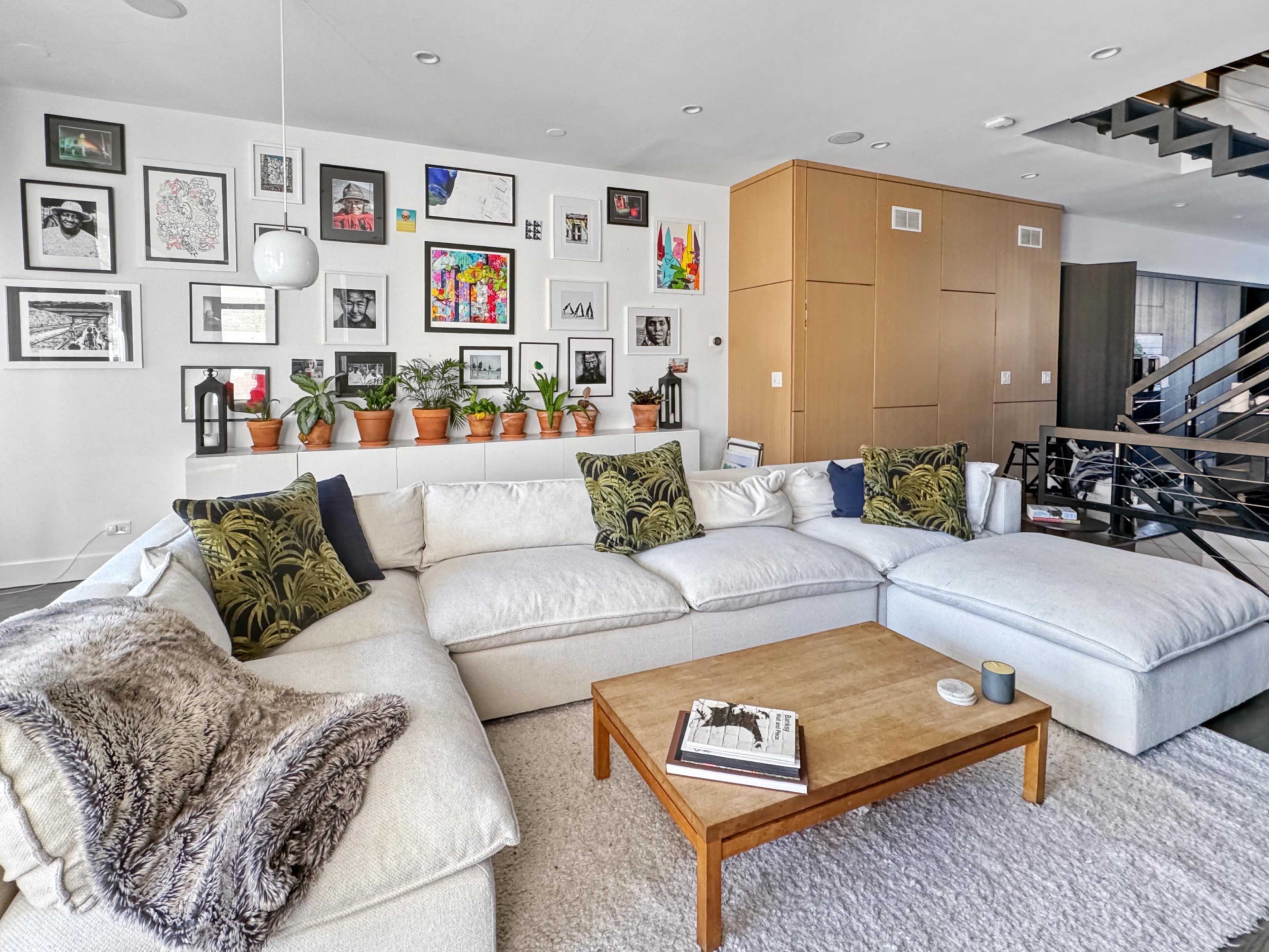 A spacious living room features a large sectional sofa adorned with patterned pillows, a wooden coffee table, and a wall decorated with framed artwork and potted plants.