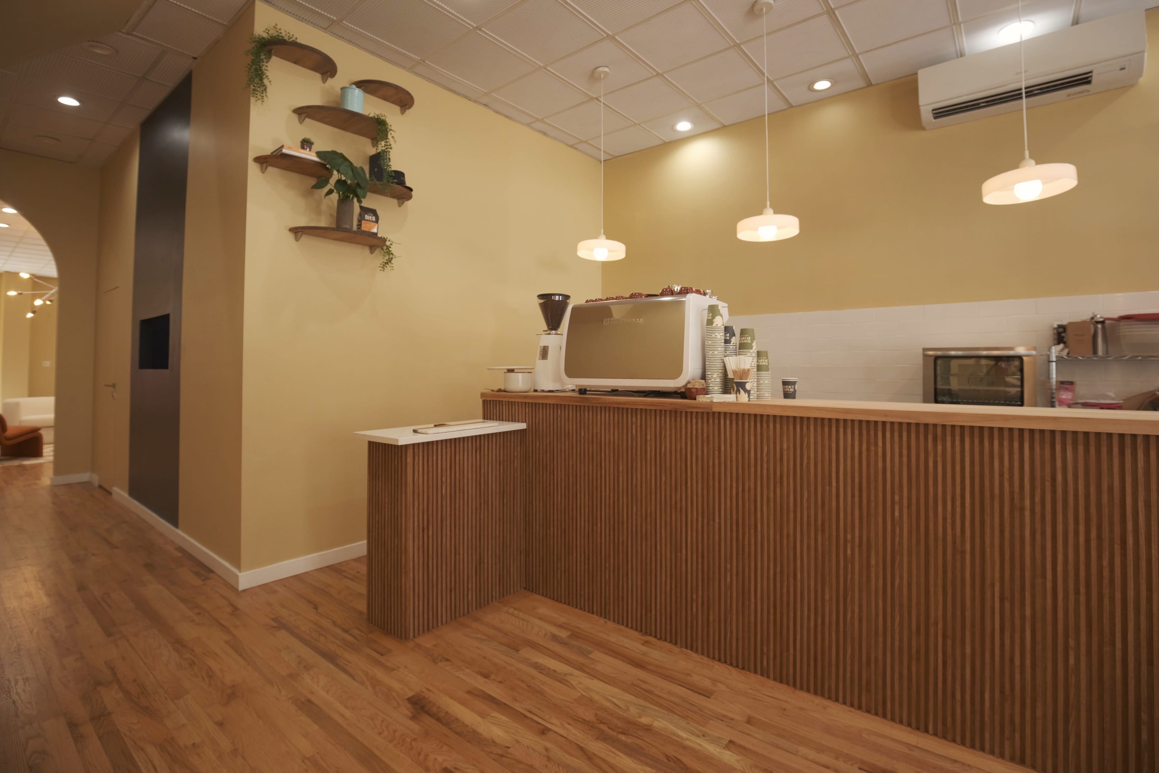A modern café interior featuring a wooden counter, a small coffee machine, and wall-mounted shelves with plants against a light-colored wall.