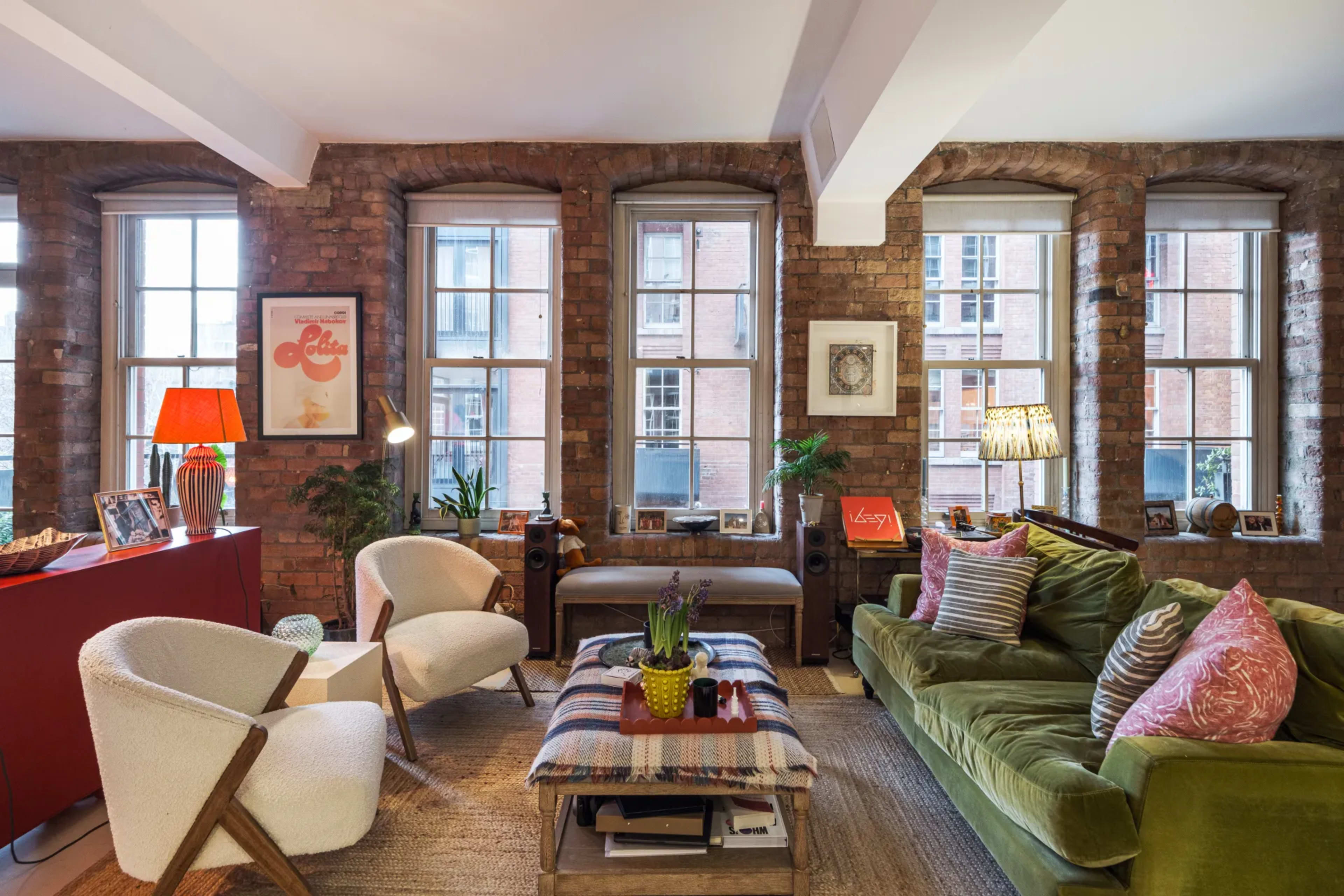 The image shows a cozy living room with exposed brick walls, large windows, and a mix of modern and vintage furniture, including a green sofa and white armchairs.