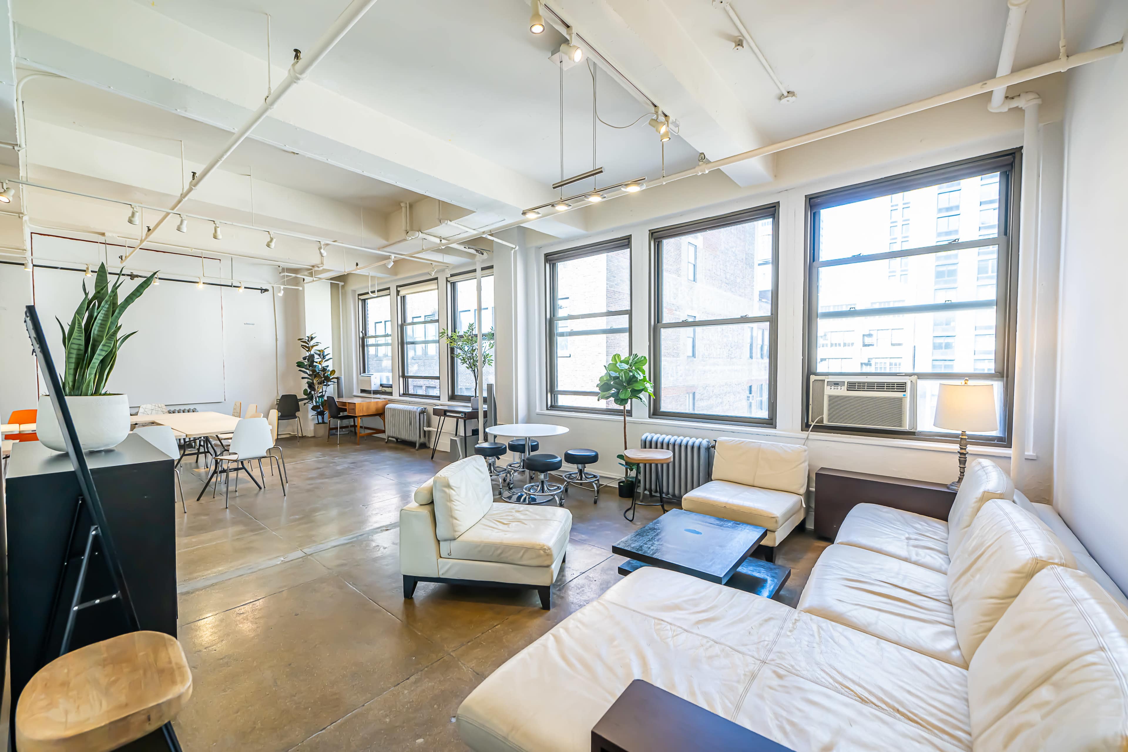 A spacious, well-lit studio apartment with large windows, featuring a mix of seating arrangements, plants, and an air conditioning unit.