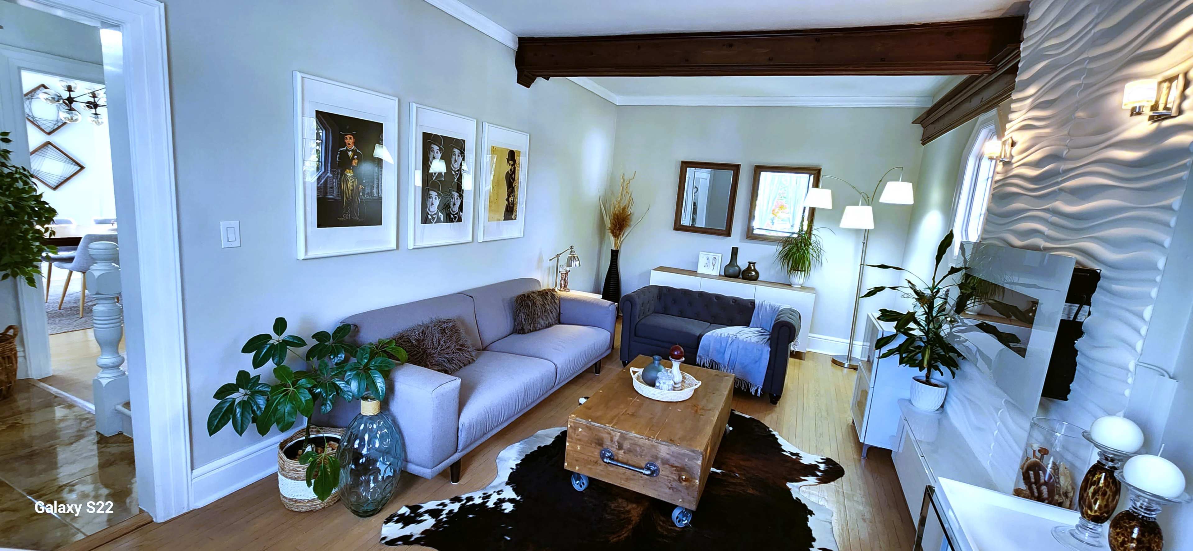 A living room with a gray sofa, a blue tufted armchair, a wooden coffee table, and decorative plants, all set against a light-colored wall with framed artwork.