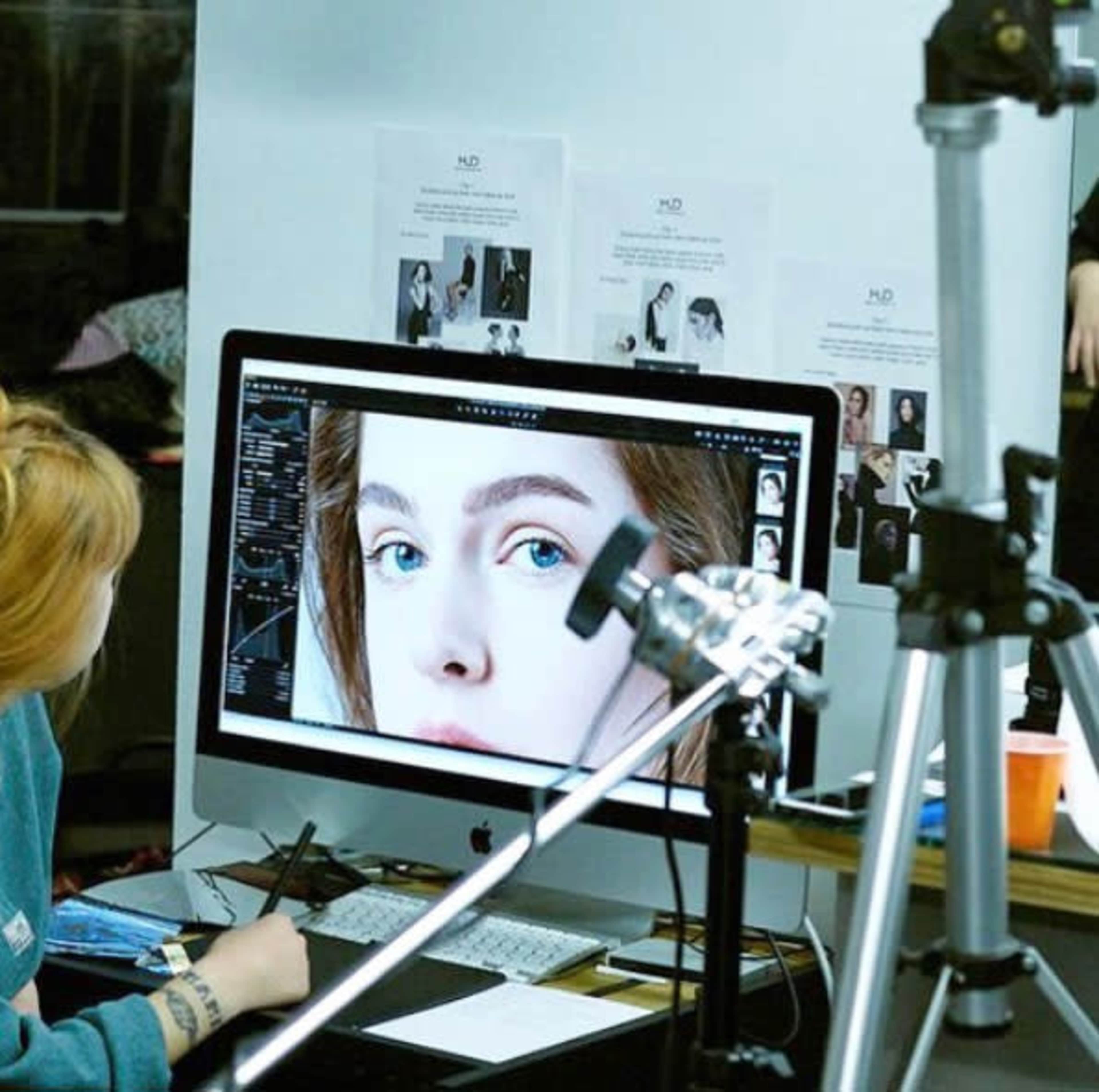 A person is seated at a desk, editing a close-up photo of a face on a computer screen, with printed reference images displayed in the background.