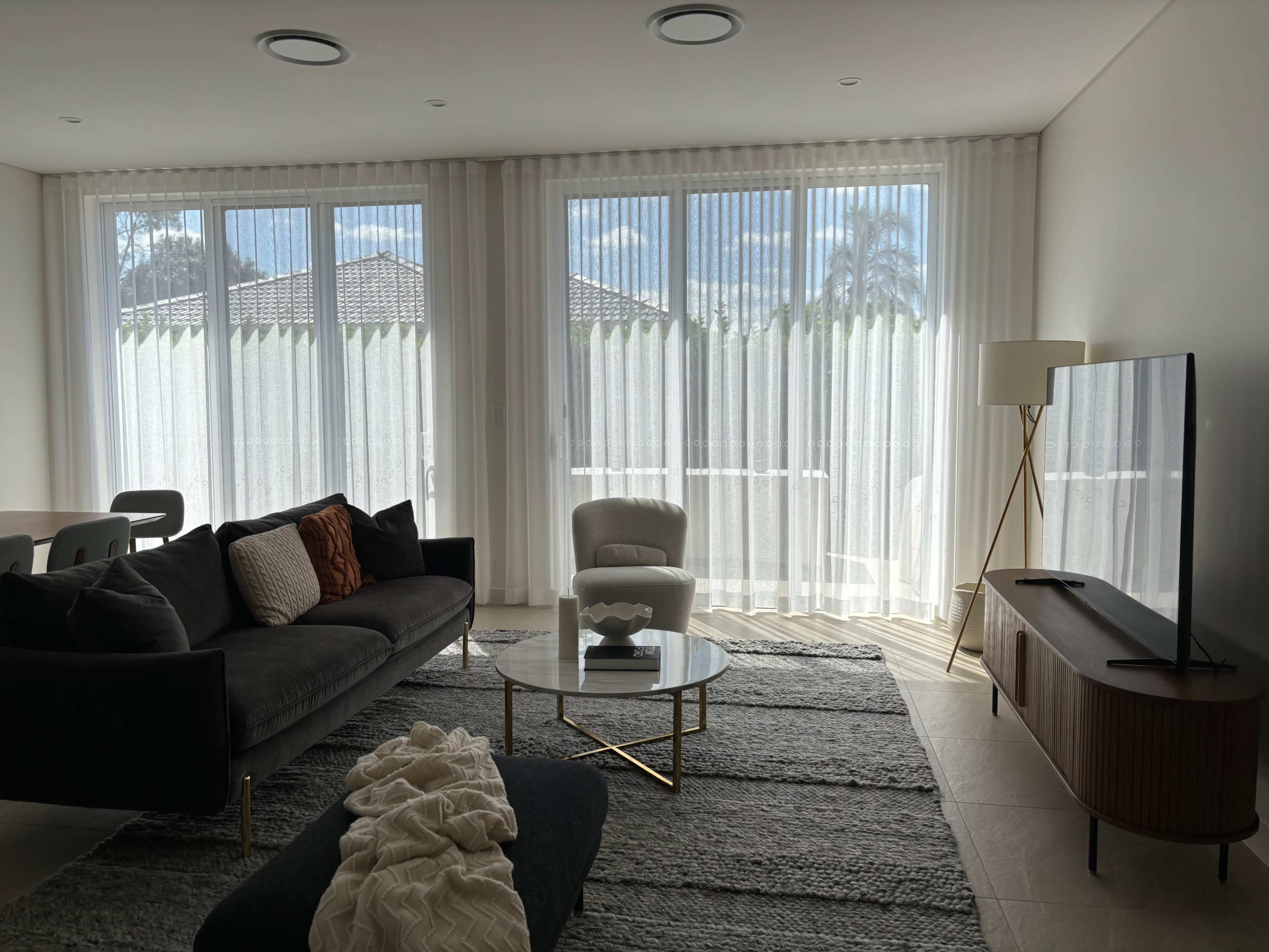 A modern living room features a dark sofa, a glass coffee table, and large windows dressed with sheer curtains, illuminating the space.