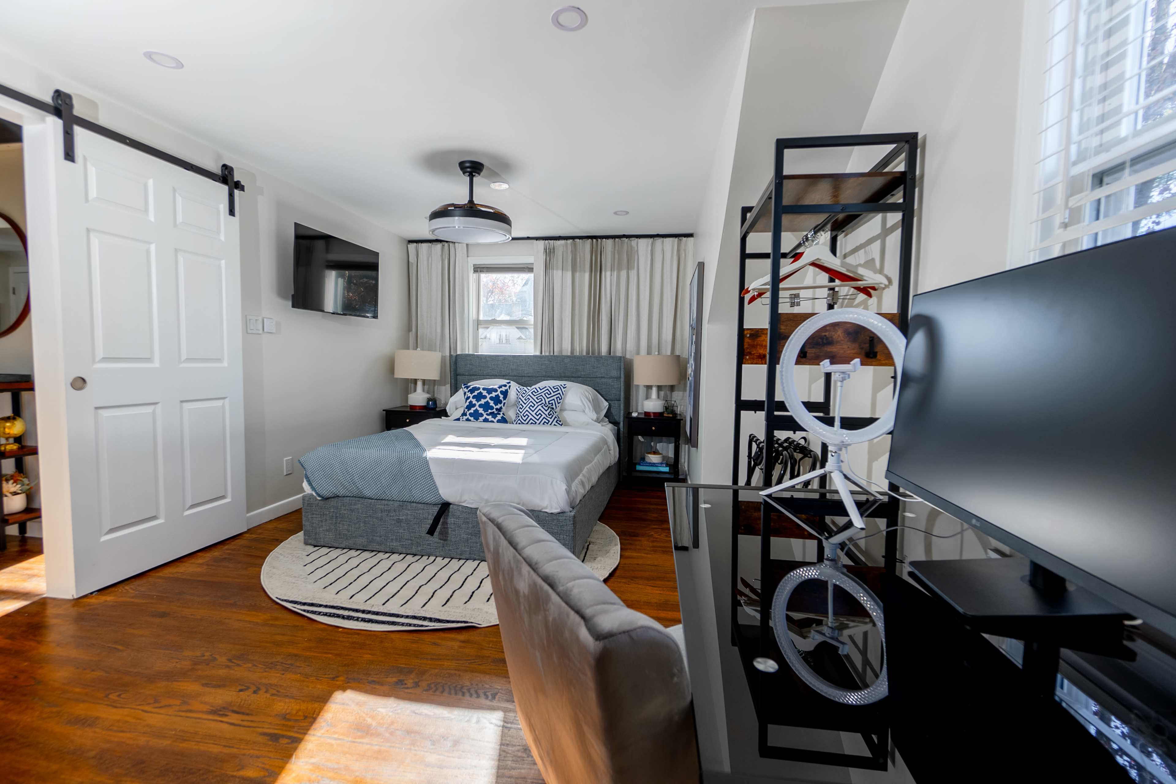 A neatly organized bedroom features a bed with a gray upholstered frame, a desk with a computer, and a large mirror, alongside a sliding door and a window with sheer curtains.