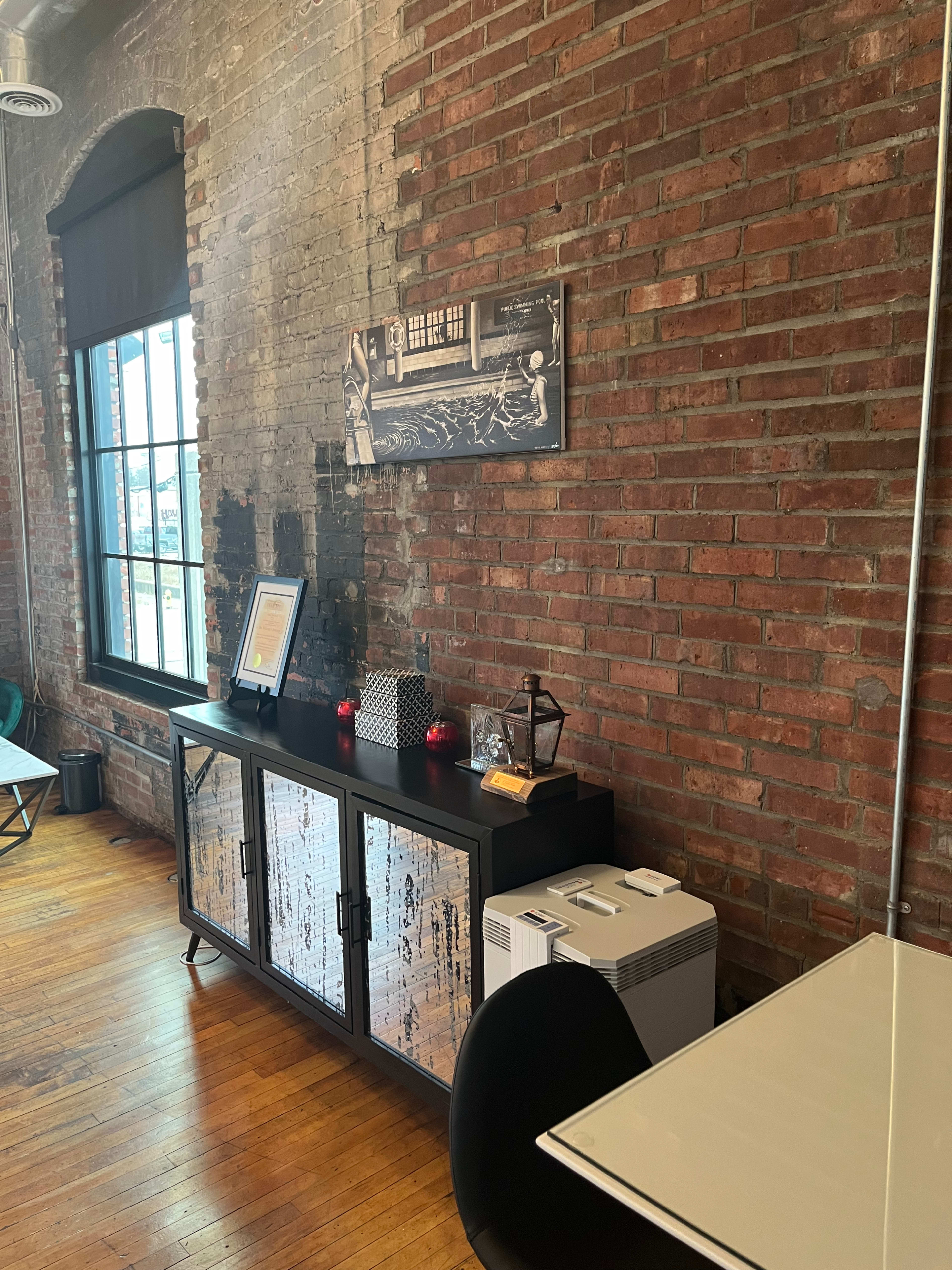 A brick wall with a window, a wooden floor, and a sideboard displaying decorative items such as candles and a framed picture.