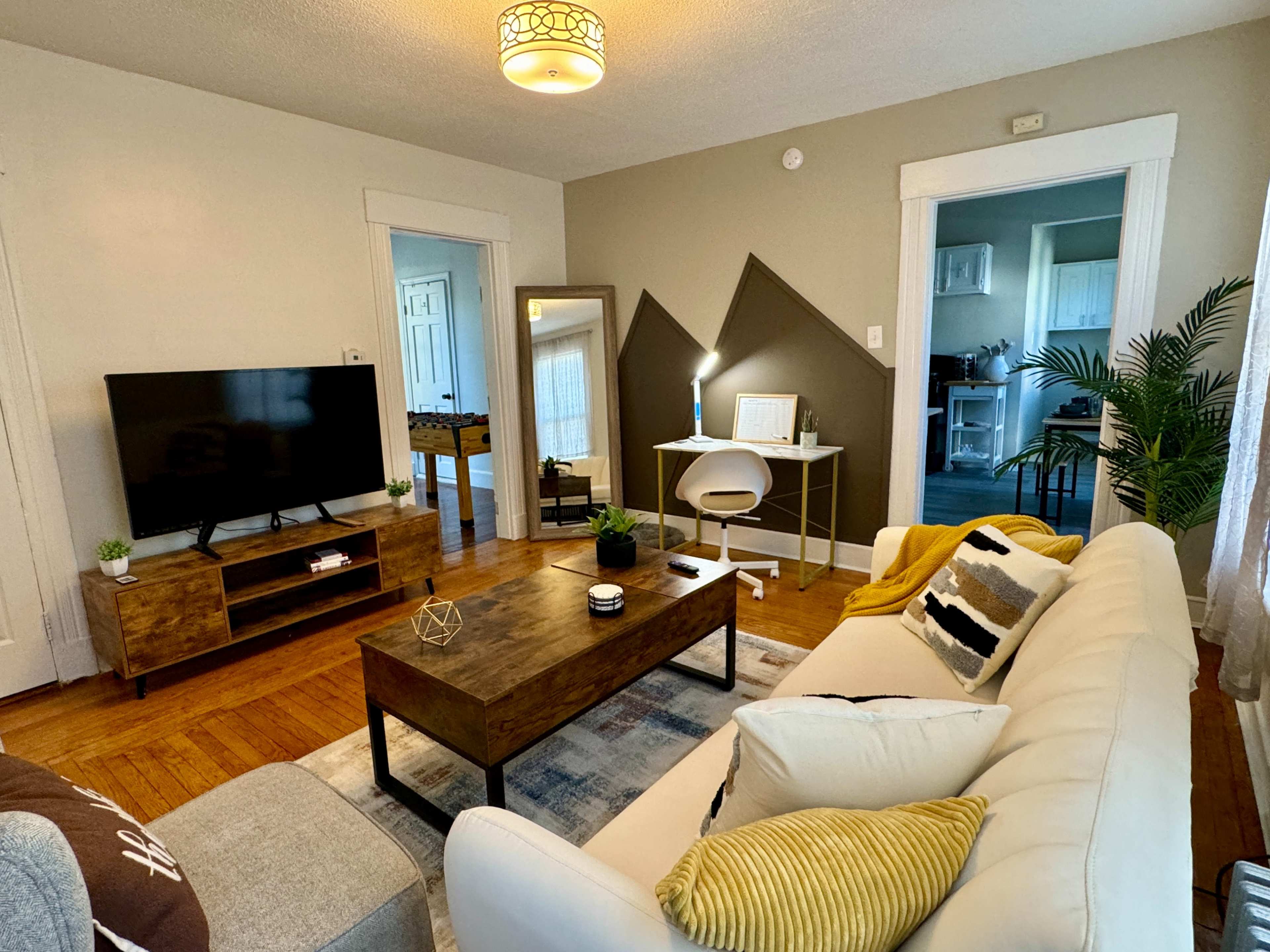 The living room features a white sofa, a wooden coffee table, a television on a low cabinet, and a workspace with a chair and lamp against a backdrop of light-colored walls and angles.