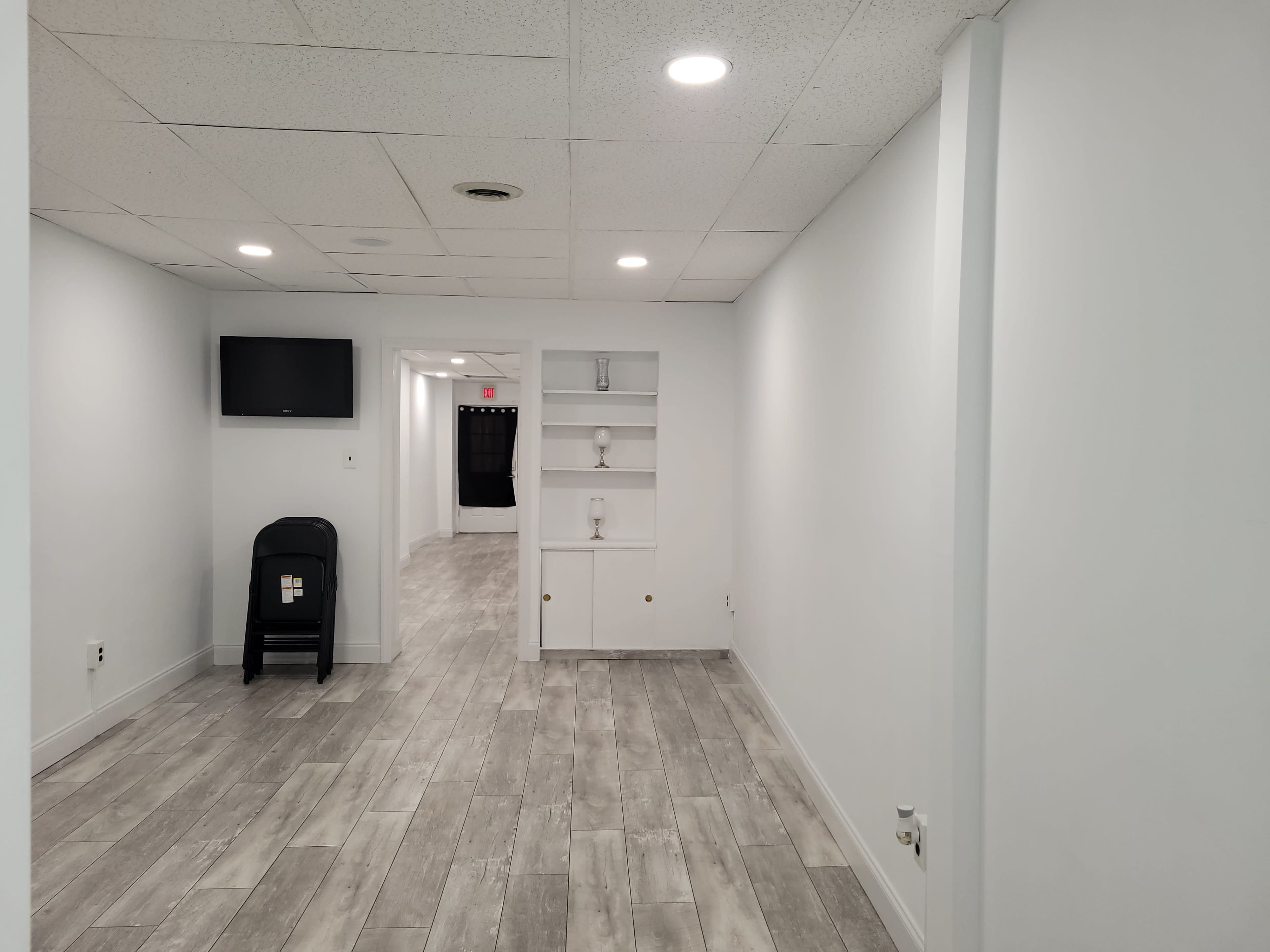 A well-lit, empty room with light-colored walls and flooring, featuring a TV mounted on the wall and a small shelf unit.