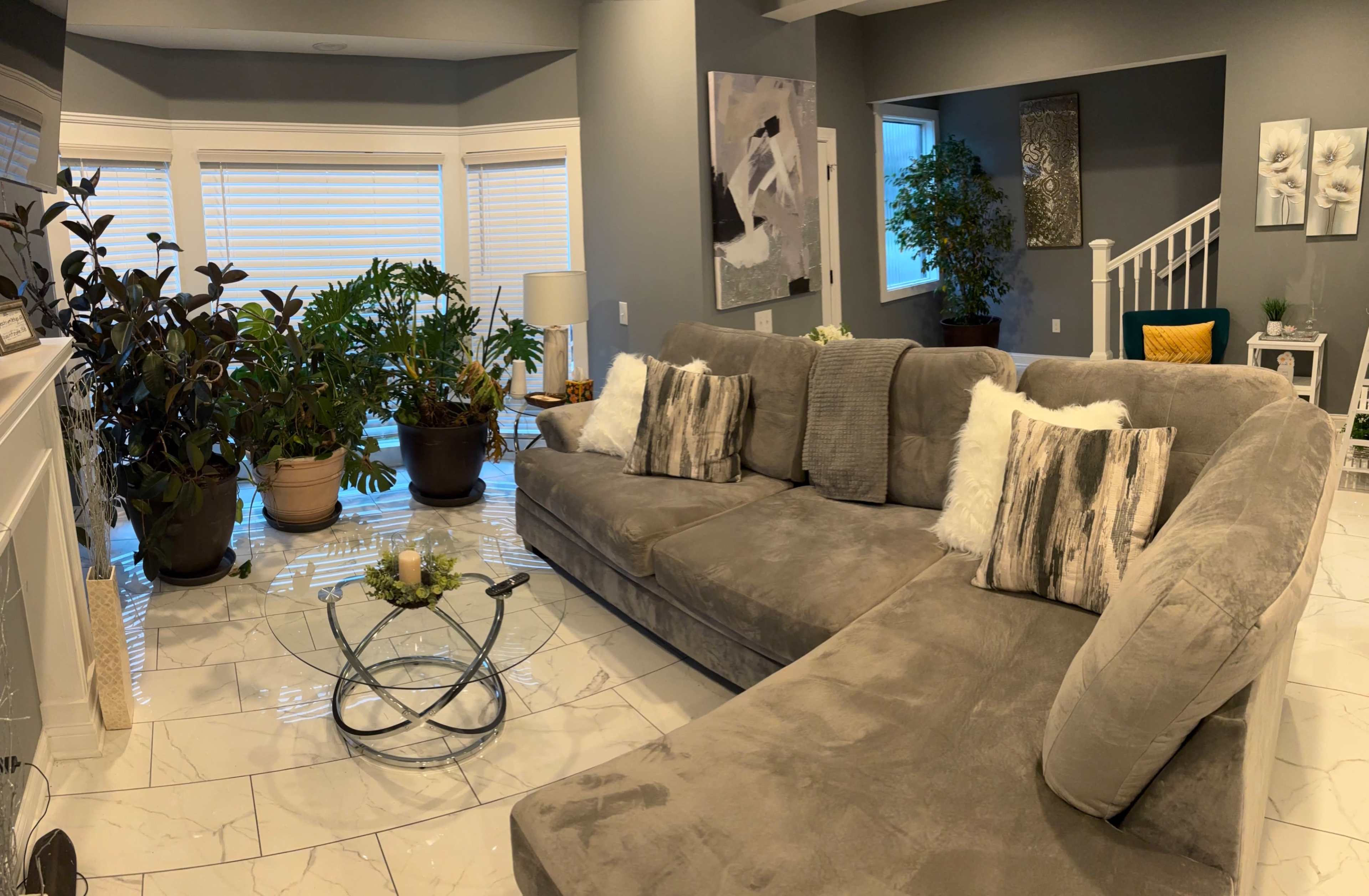 The image shows a modern living room with a large gray sectional sofa, a glass coffee table, and several potted plants, all arranged around large windows that let in natural light.