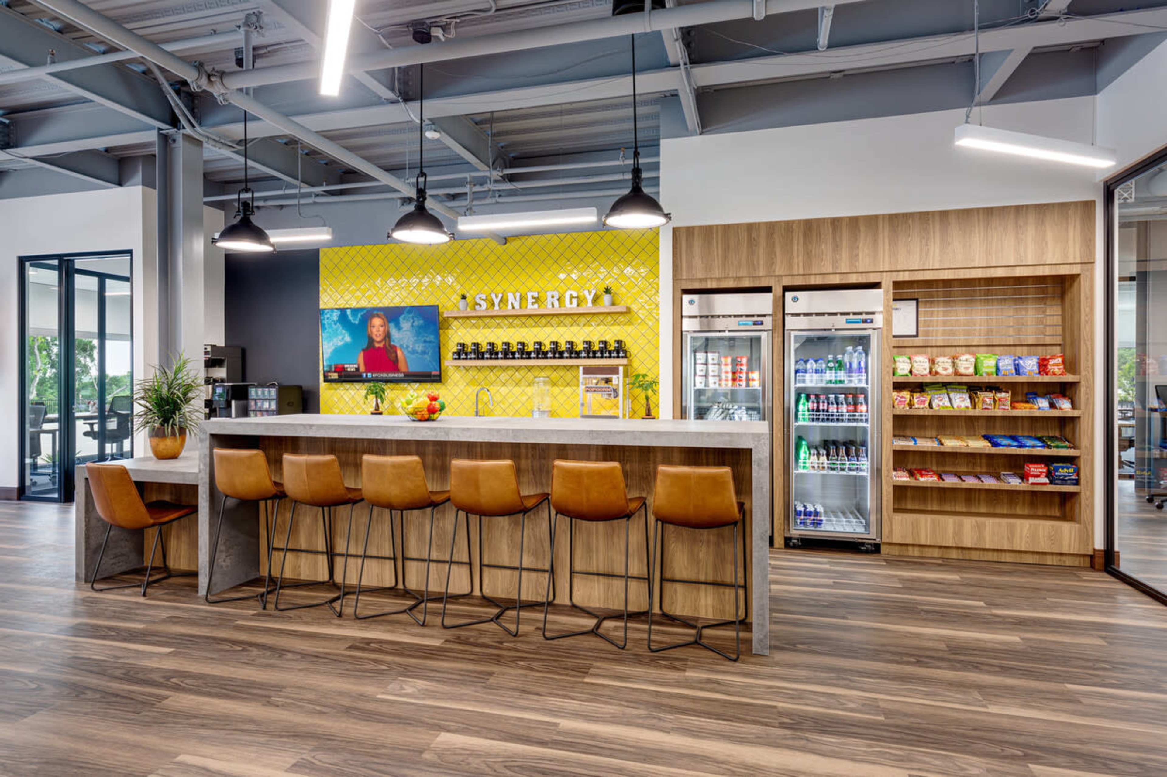 A modern office break room featuring a long bar-style seating area, a colorful accent wall with "SYNERGY" written prominently, and a self-service snack and beverage station.