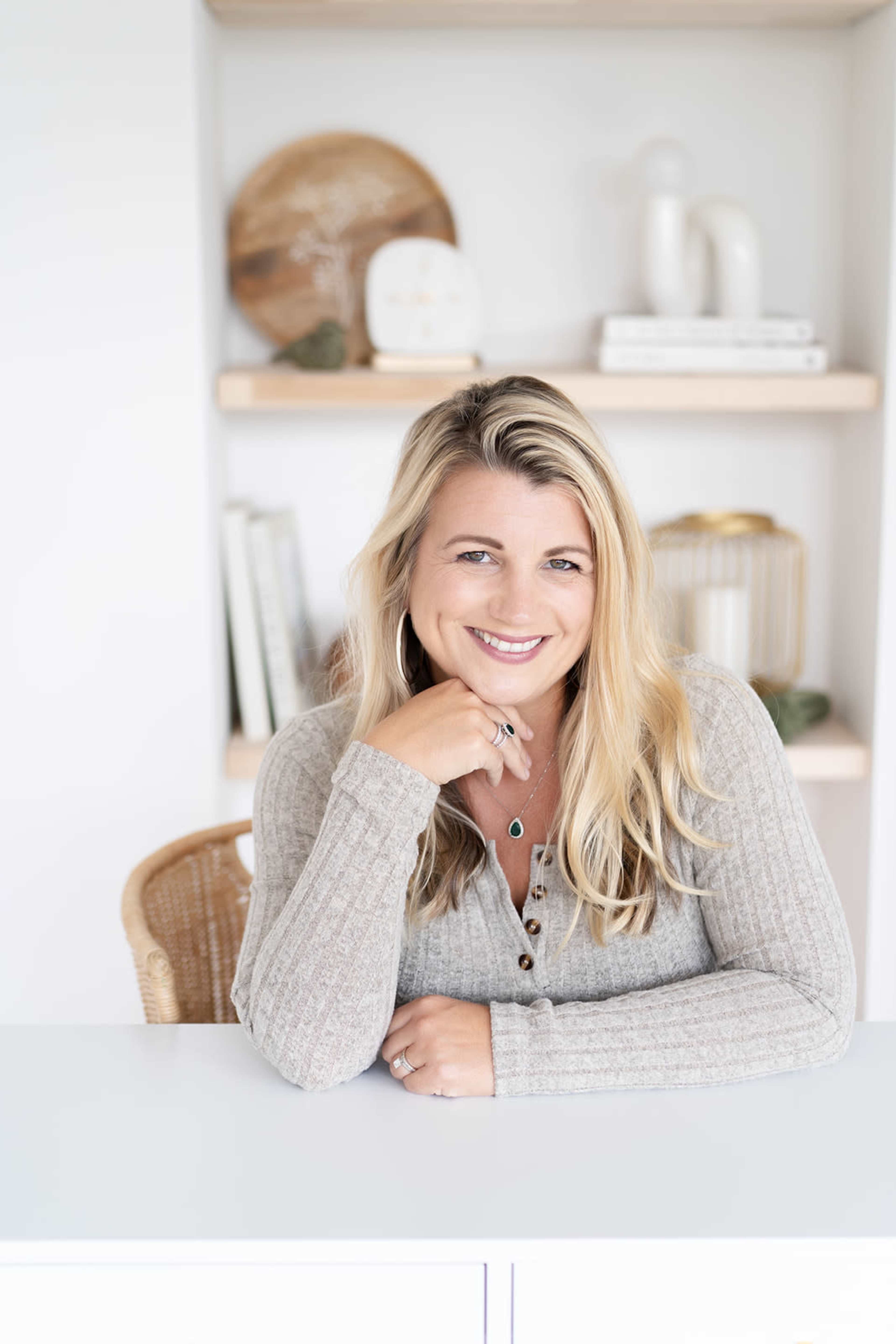A woman with long, blonde hair sits at a white table, leaning on her hand and smiling, with shelves of decor behind her.