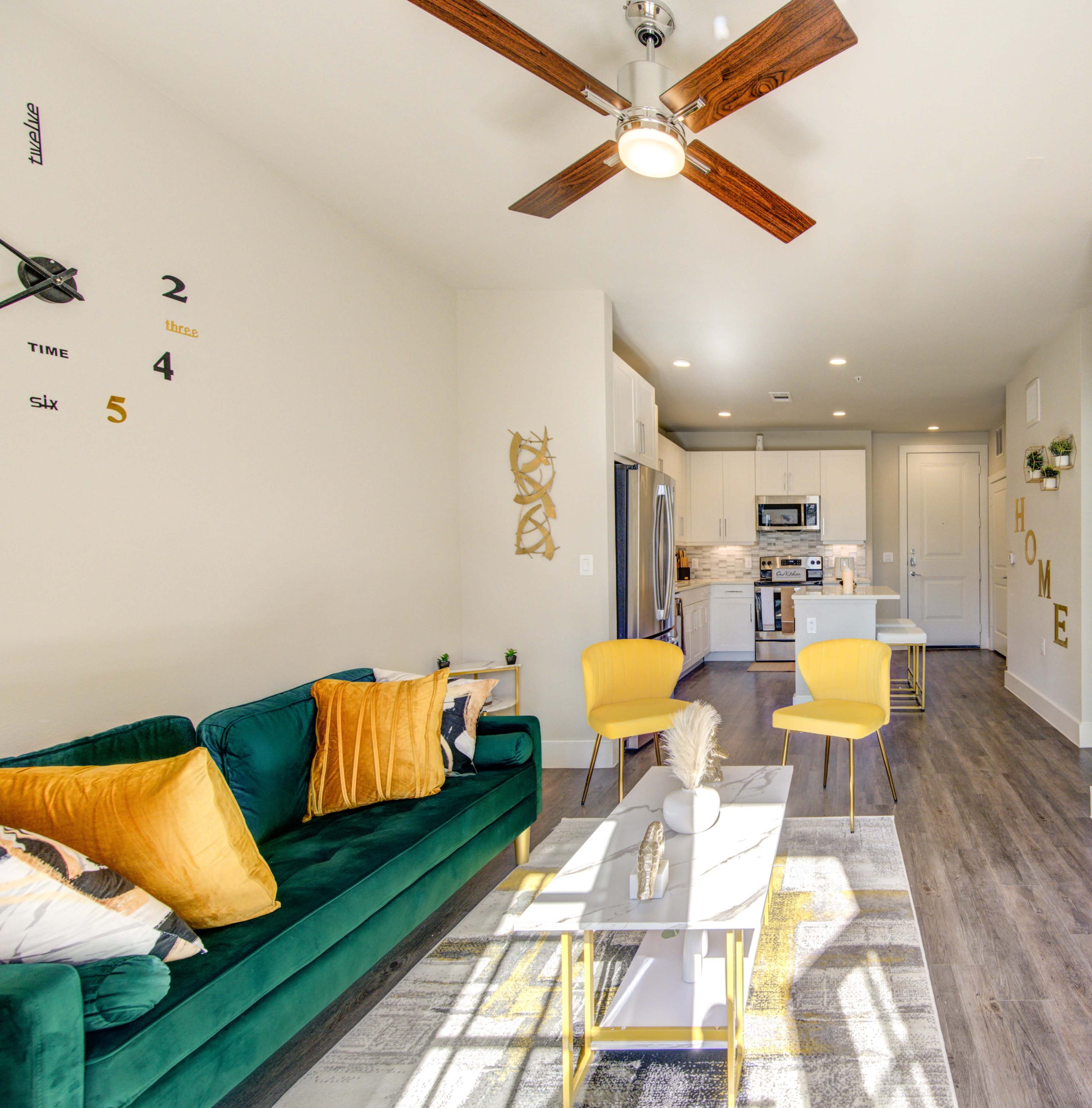 The image shows a modern living room featuring a green sofa with yellow pillows, a white coffee table, and yellow chairs, set against a light-colored wall and a kitchen area in the background.