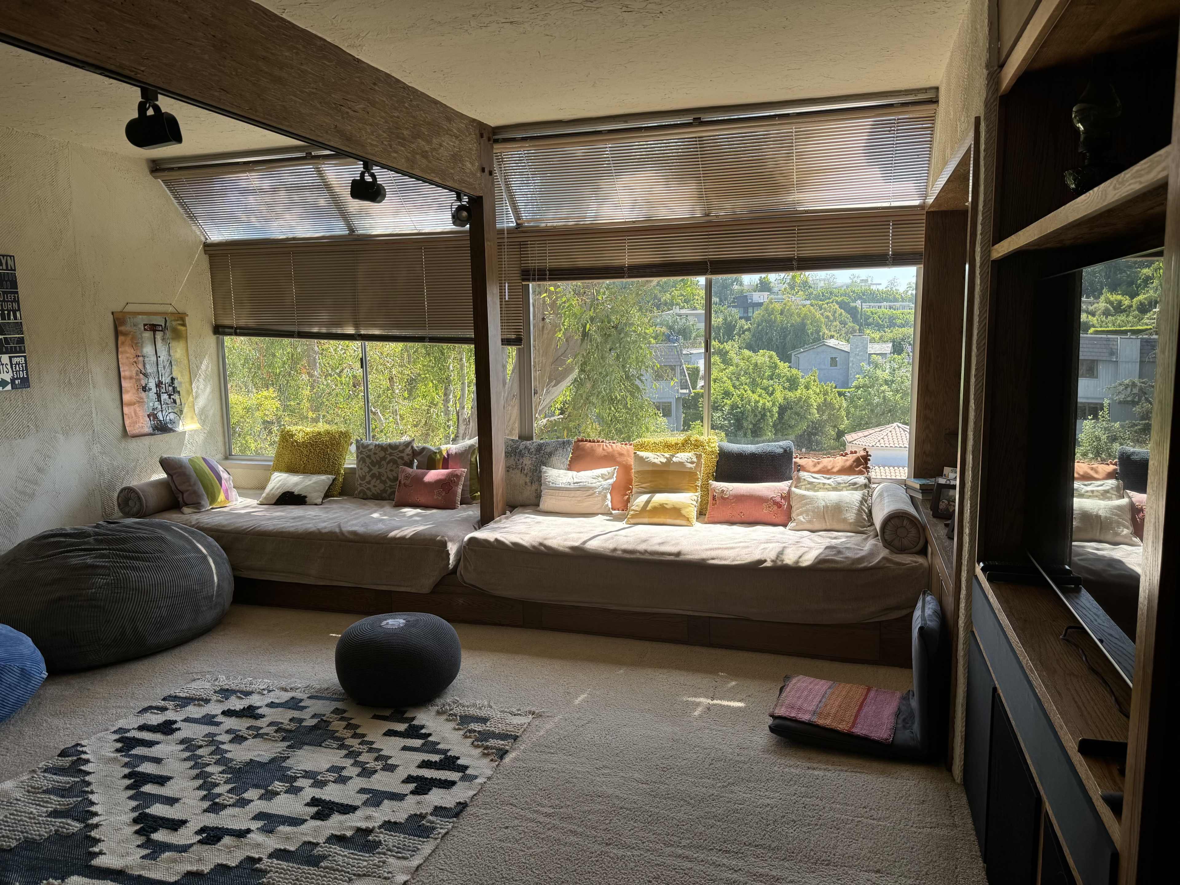 A cozy living area with large windows, featuring a low seating area adorned with colorful pillows and a patterned rug on a carpeted floor.