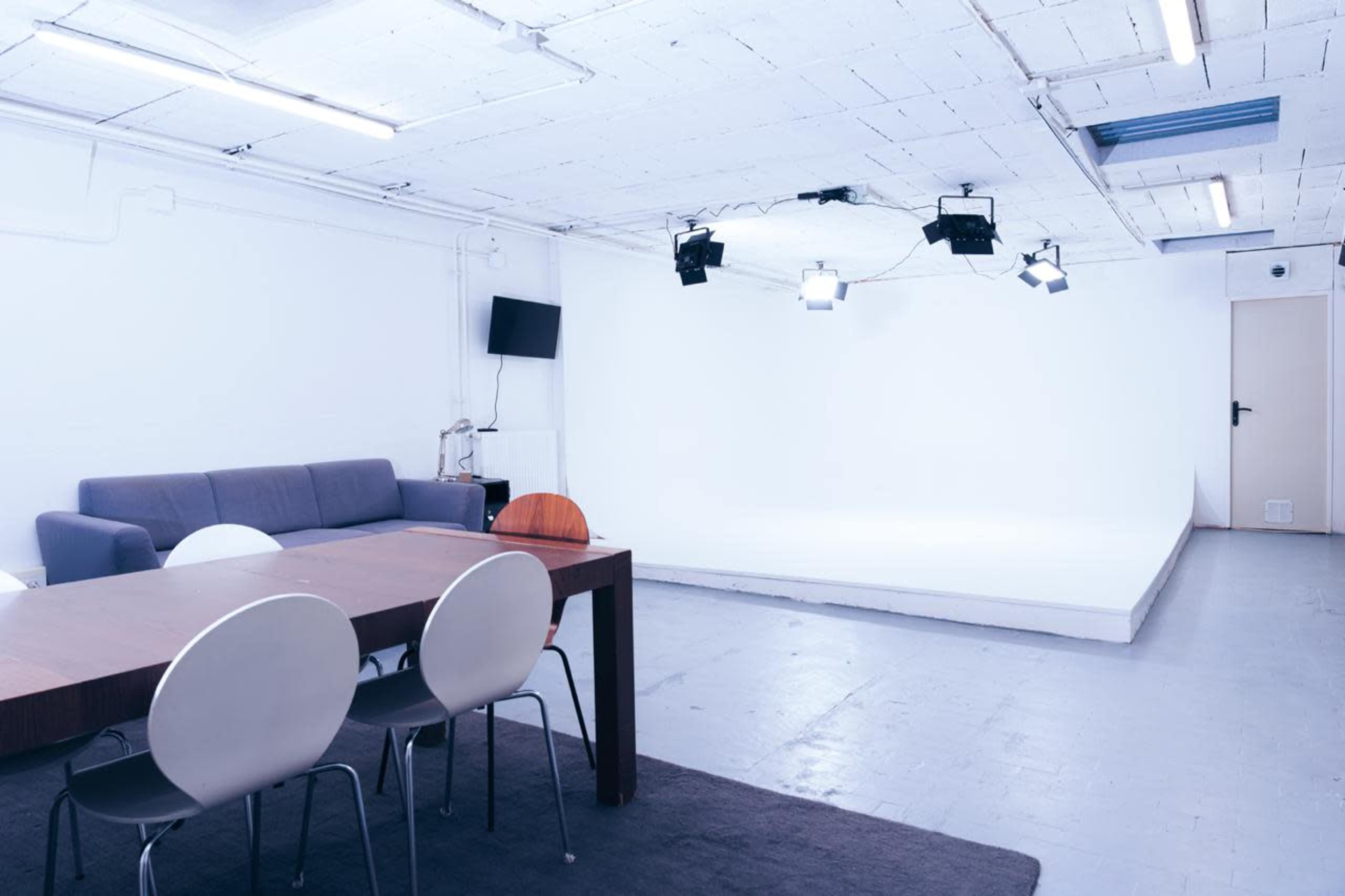 The image shows a minimalist studio space featuring a grey couch, a wooden dining table with white chairs, and a bright, empty stage area illuminated by overhead lights.