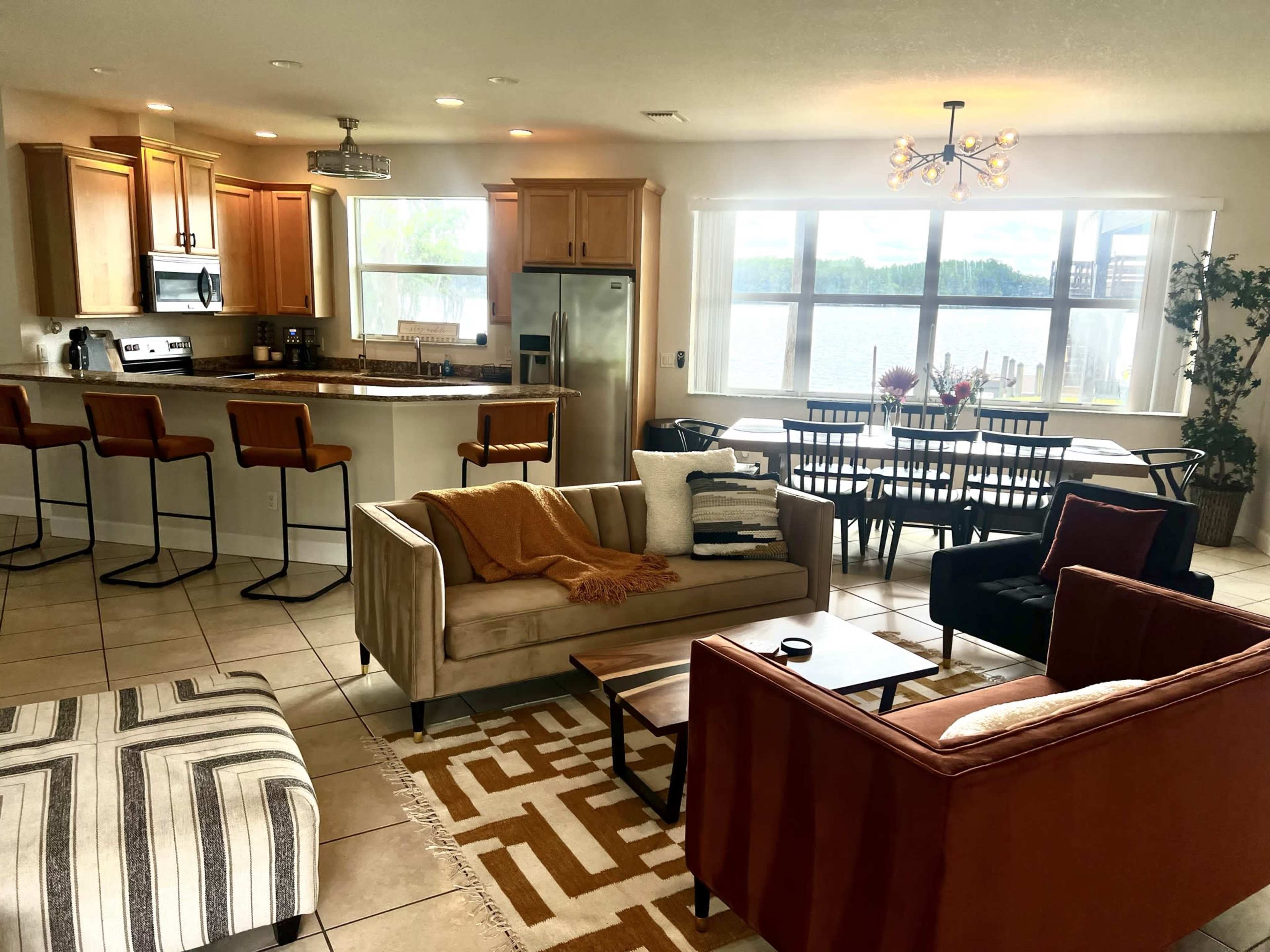A modern open-concept living area with a kitchen, dining table, and seating arrangements, all overlooking a lake through large windows.