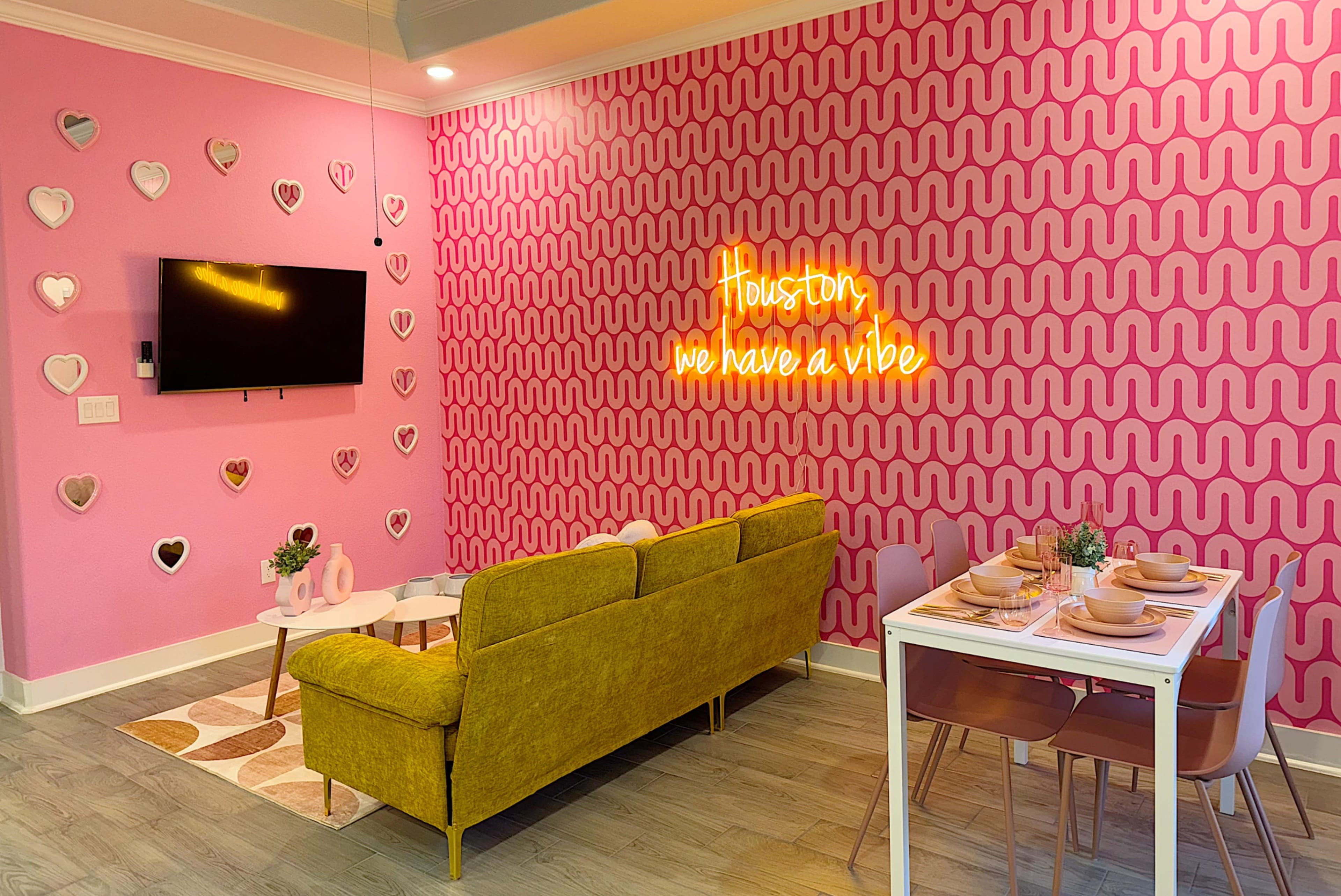 A modern living space with a pink patterned wall, a yellow sofa, a television, and a dining table set for four.