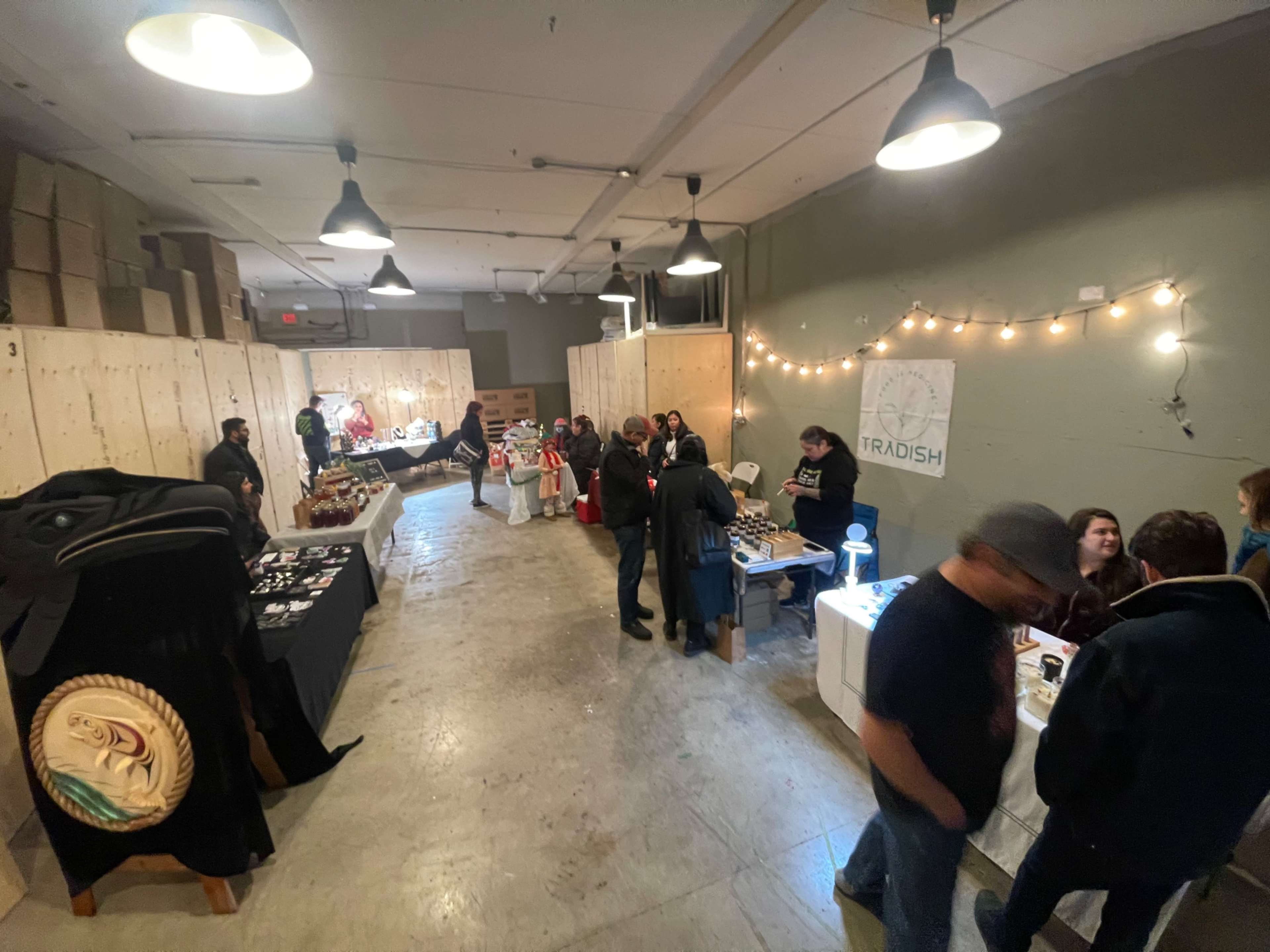 A crowded indoor marketplace features various tables displaying products, with shoppers interacting and browsing among the stalls.