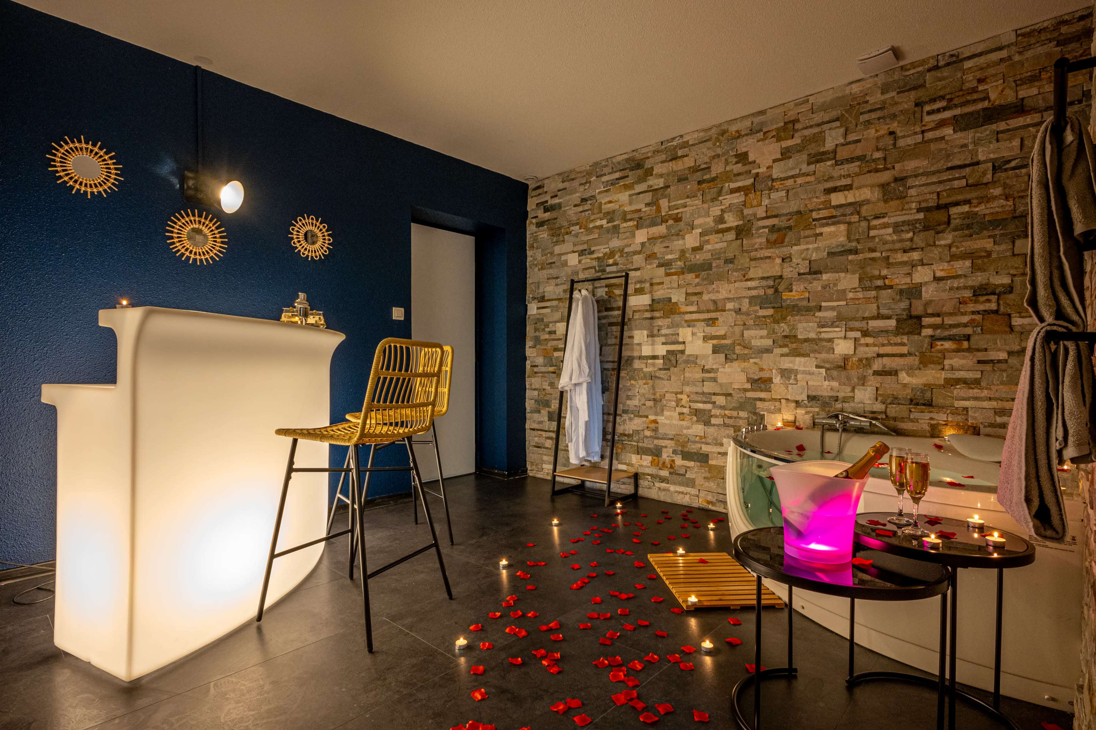 A modern bathroom with a stone wall, a glowing white bar counter, a yellow chair, a bathtub surrounded by candles, and rose petals scattered on the floor.