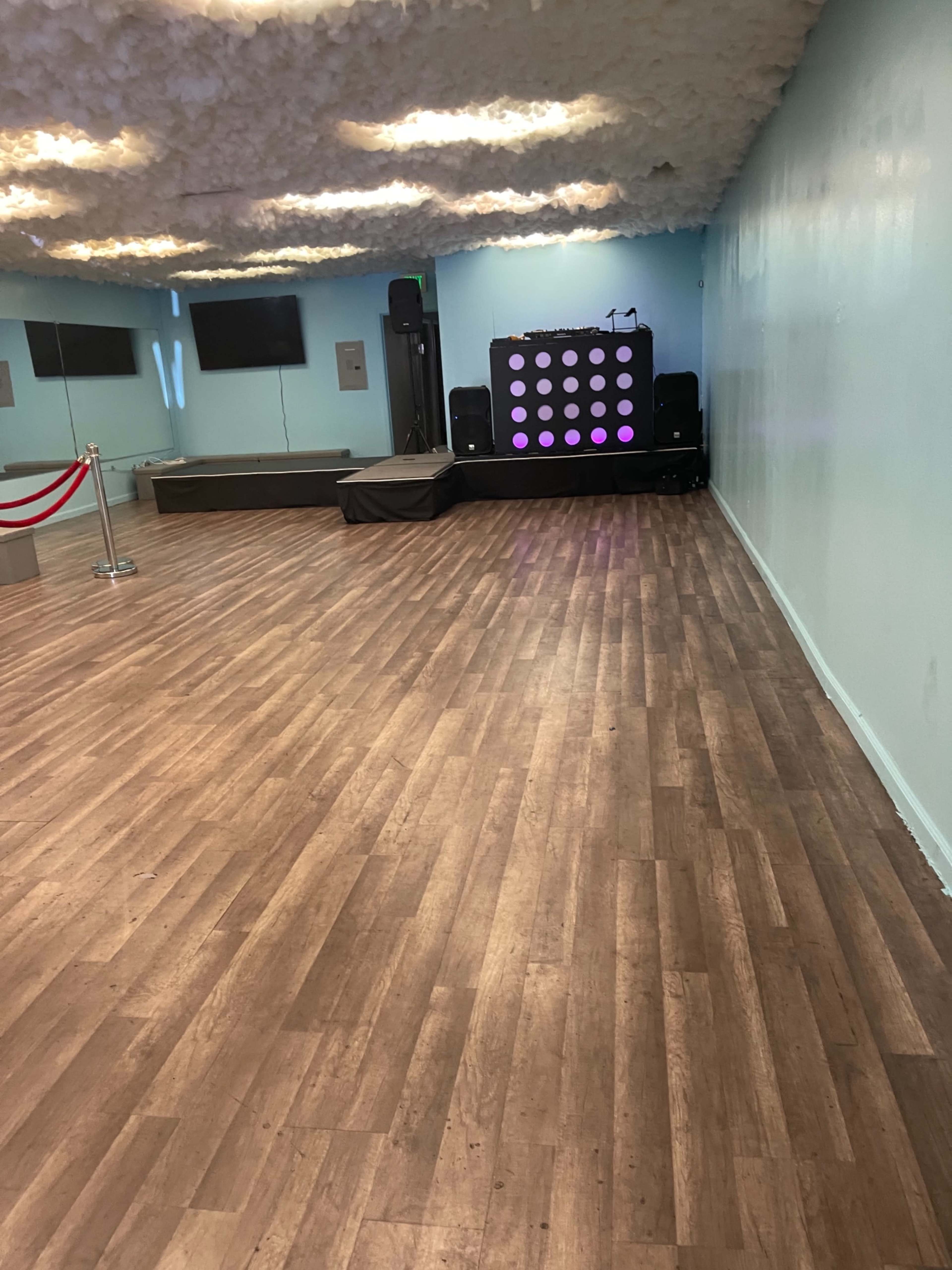 A spacious event room with wooden flooring, a stage at the back featuring a music setup, and soft, cloud-like ceiling decorations.