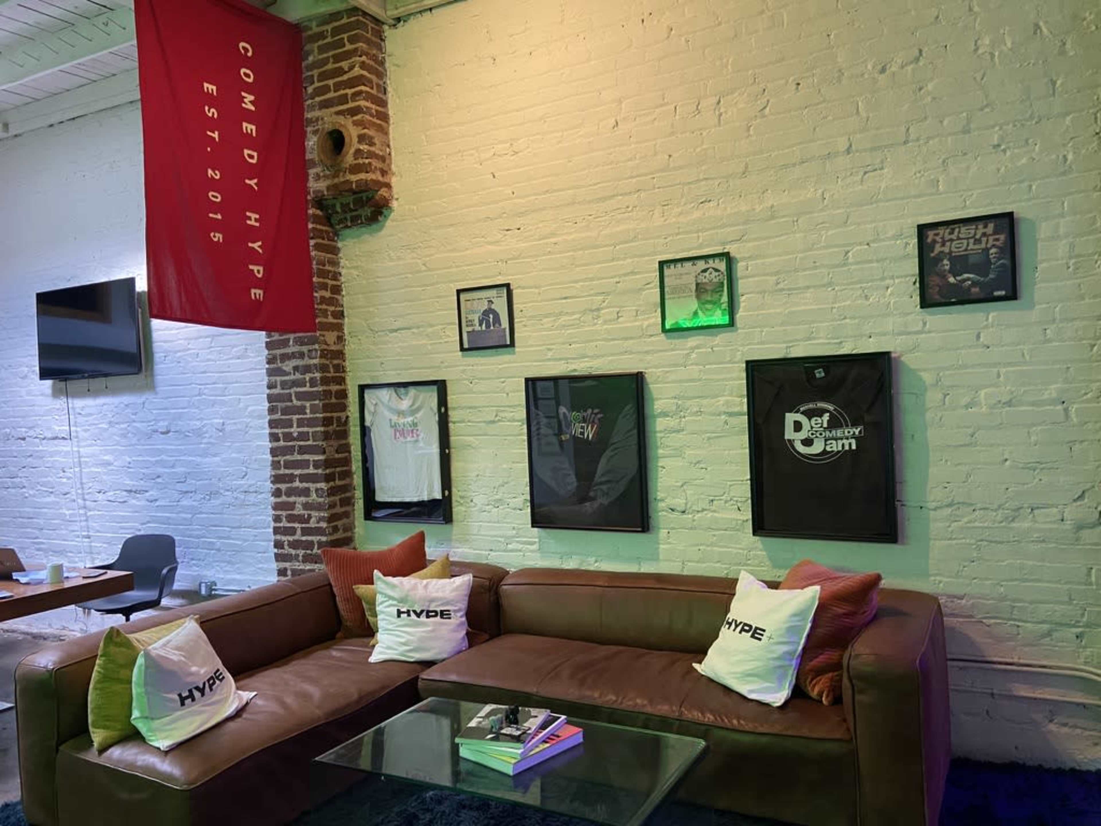 A cozy lounge area with a brown leather couch, decorative pillows, and a wall displaying framed posters and a banner for "Comedy Hype."