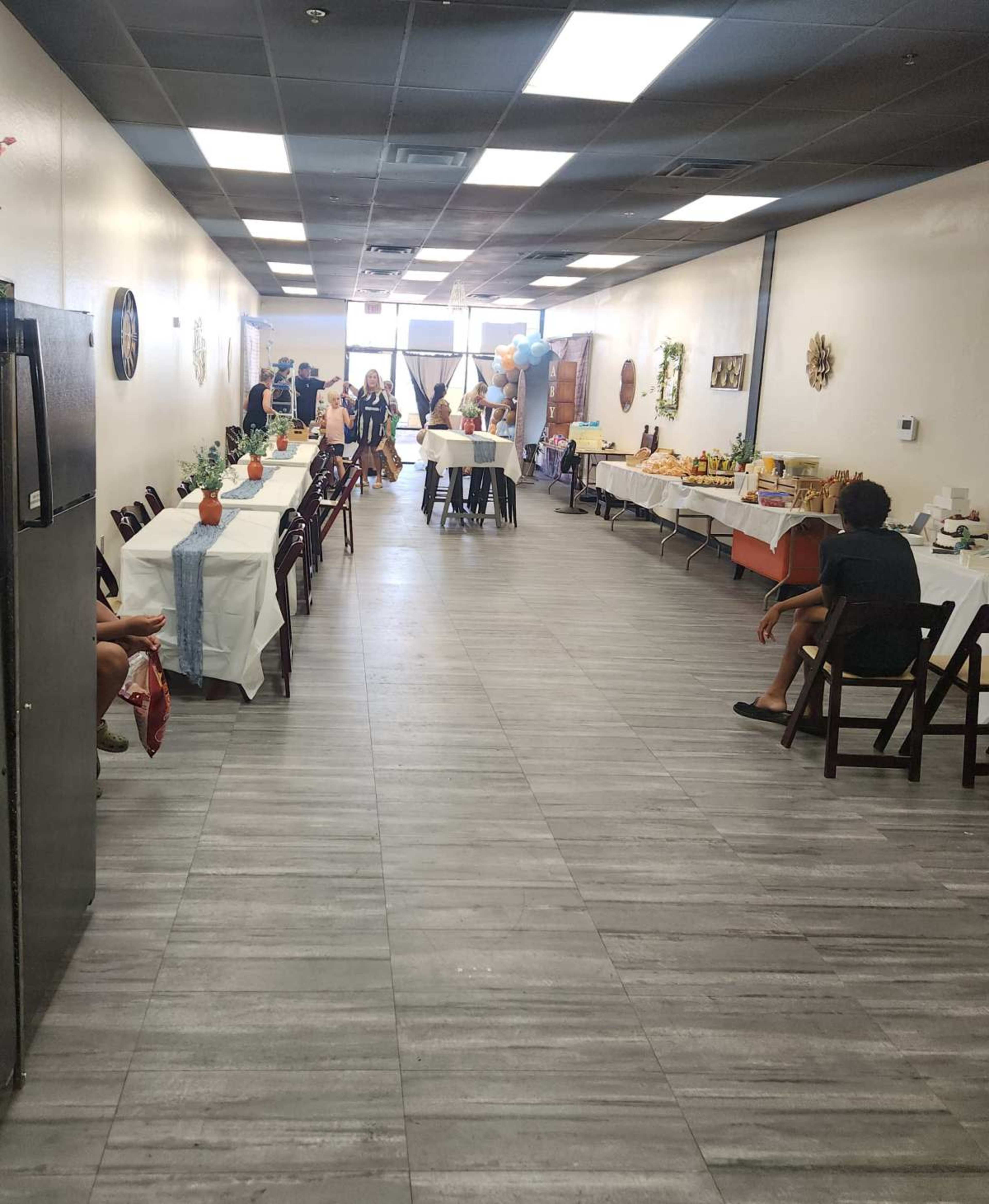 The image shows a long, narrow room set up for an event, with tables on both sides and people in the background mingling and arranging food.