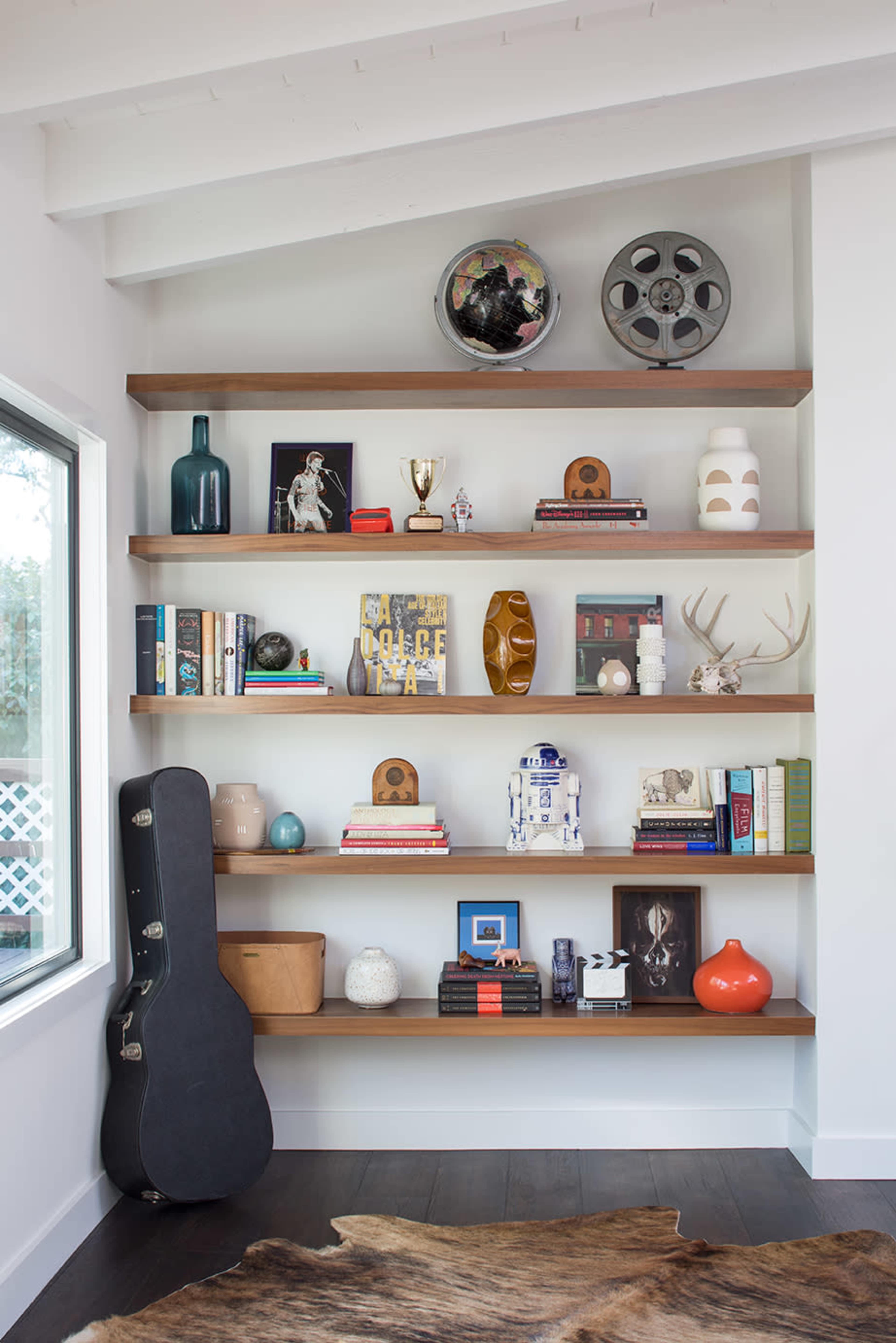 The image shows a modern shelving unit with various decorative items, books, and a guitar case arranged on wooden shelves.