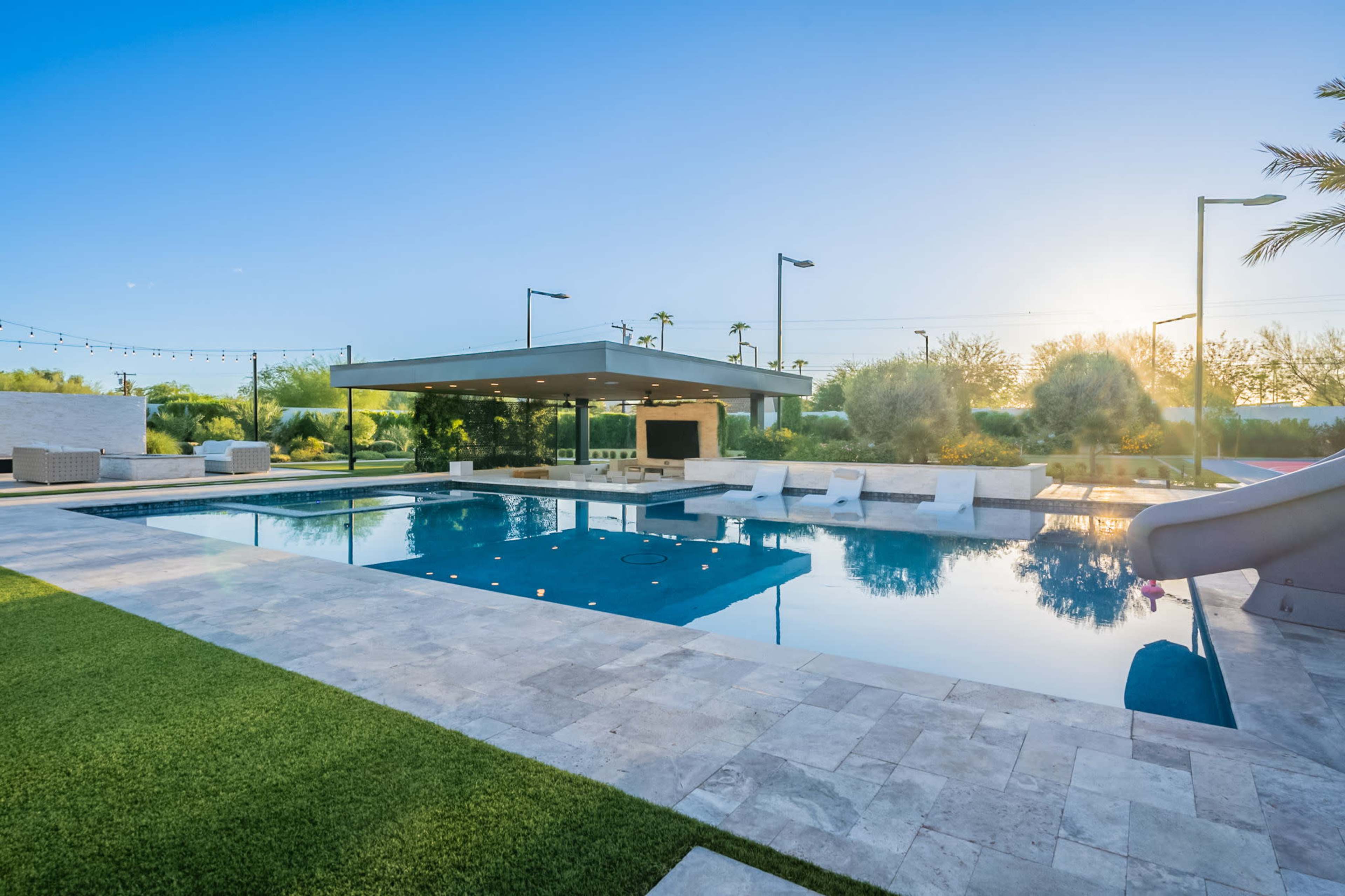 The image shows a modern outdoor swimming pool with a water slide, surrounded by a grassy area and landscaped gardens, under a clear blue sky.