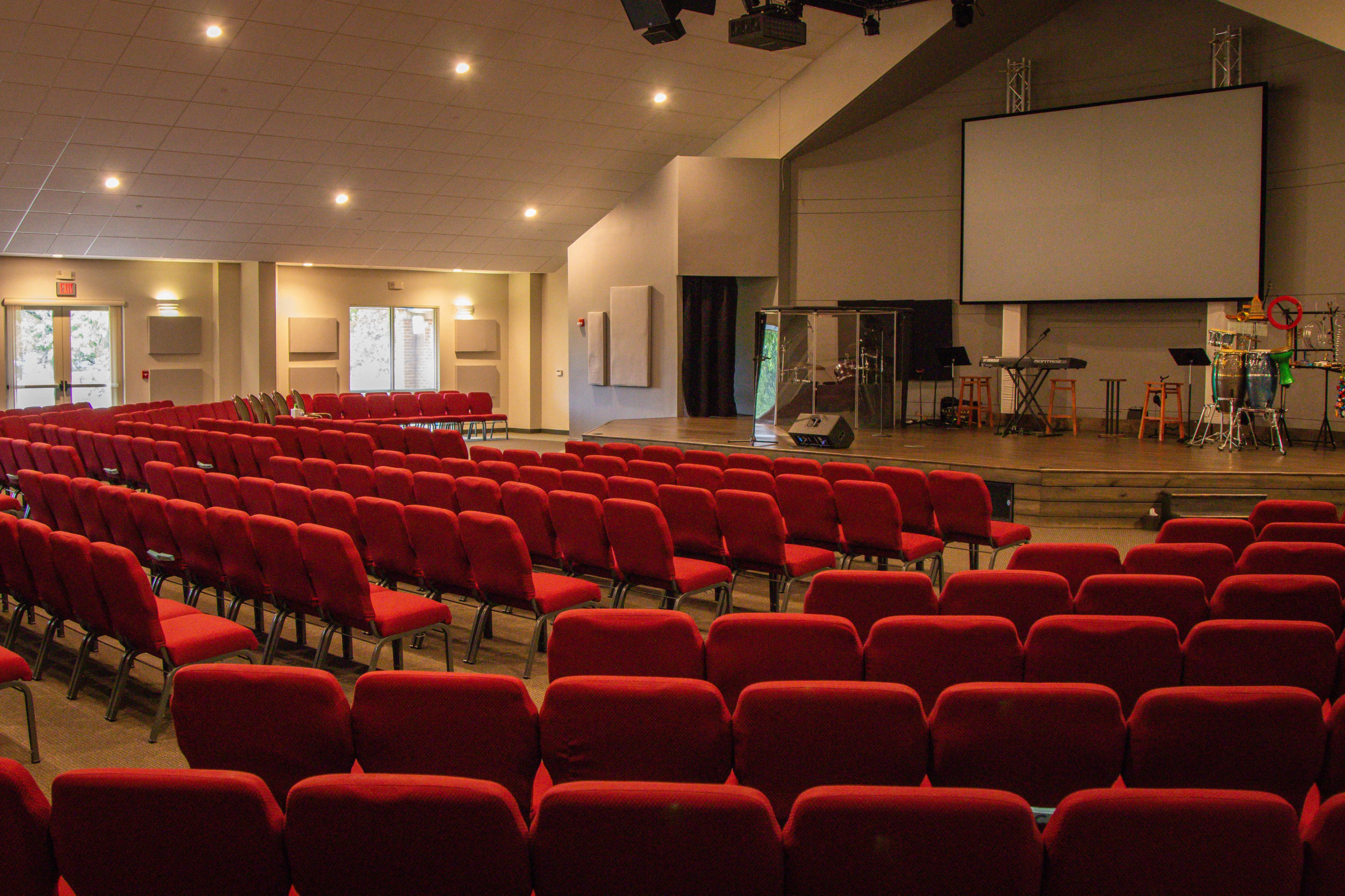 A spacious auditorium with rows of red chairs facing a stage that features a large blank screen and various musical instruments.