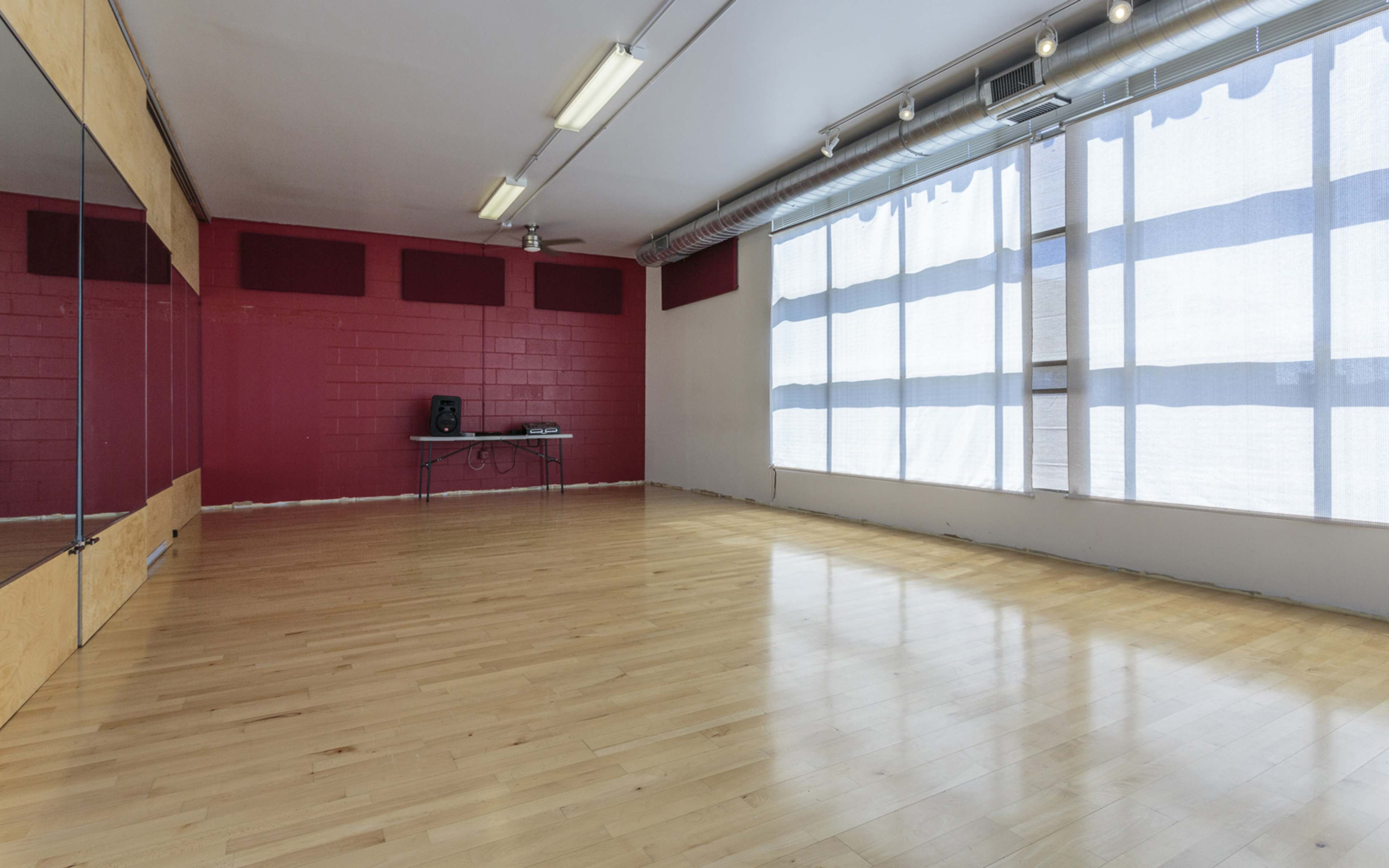 An empty dance studio with hardwood floors, red walls, a large window covered by sheer curtains, and a table with equipment against the back wall.