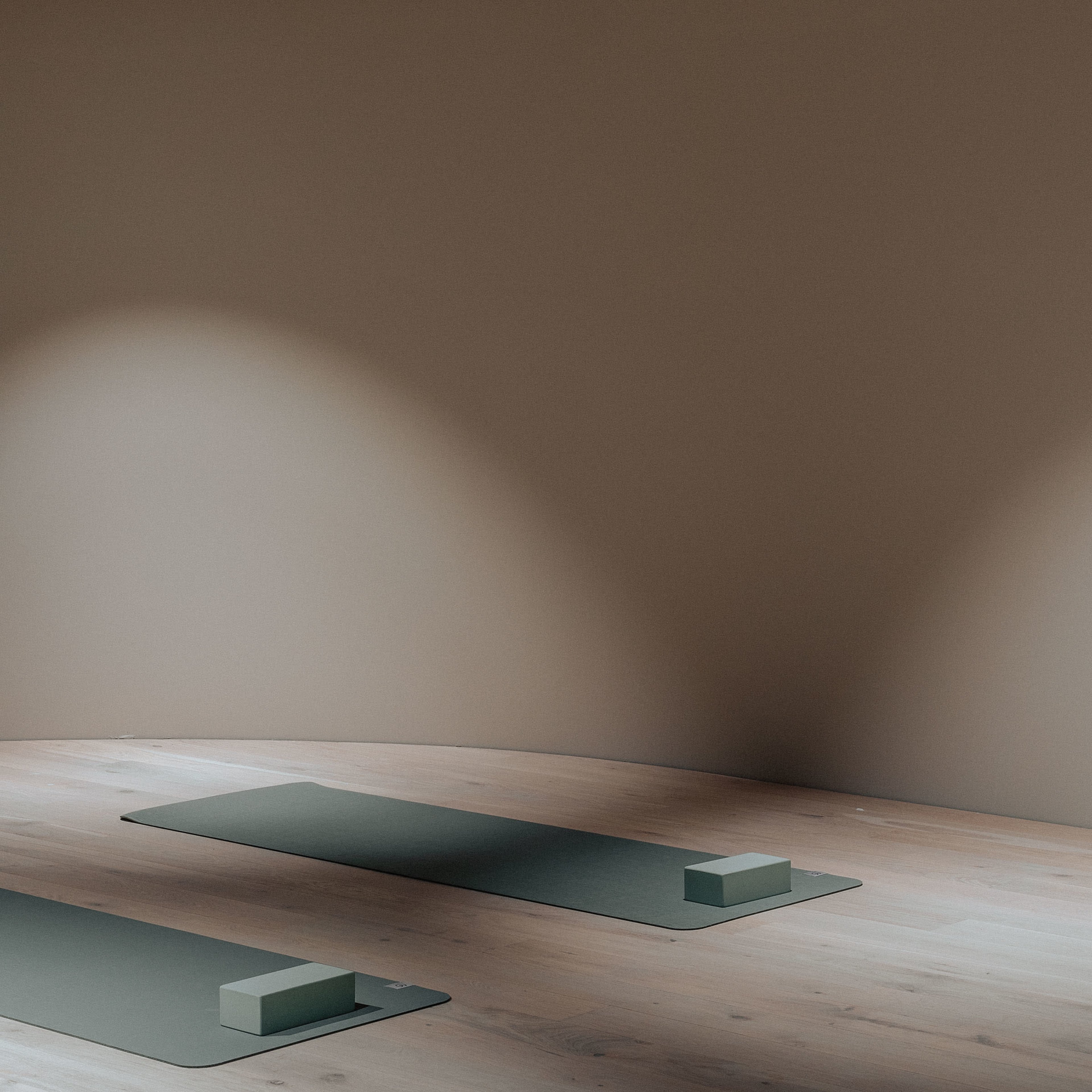 The image shows two yoga mats placed on a wooden floor in a dimly lit room with a brown wall.