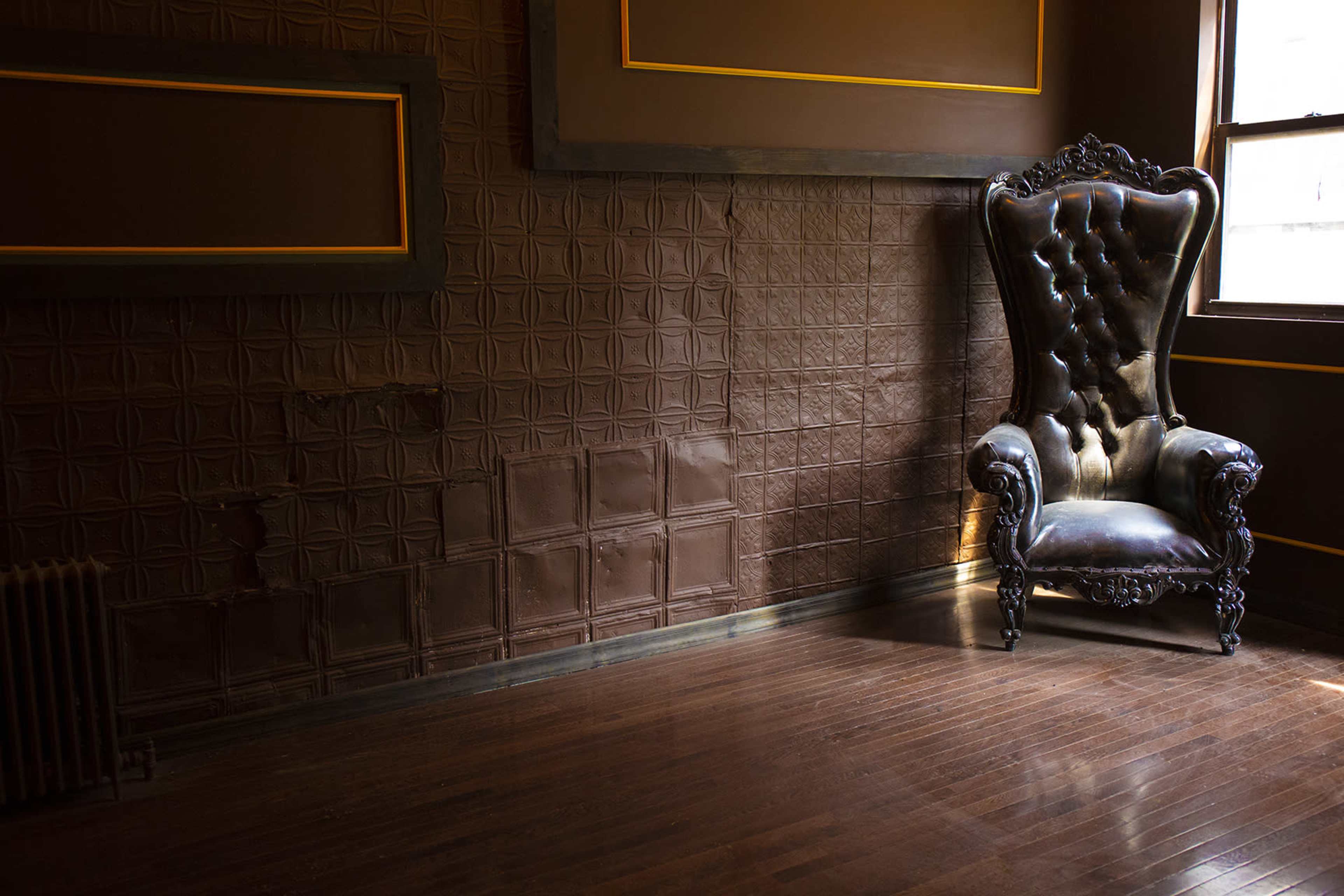 A large, ornate black chair is positioned against a wall with patterned panels and gold trim in a dimly lit room.